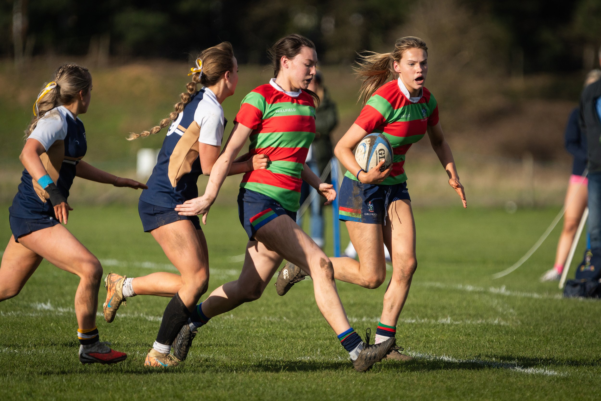 Girls rugby players competing on a grassy field during a match, with some wearing striped uniforms and holding a rugby ball.