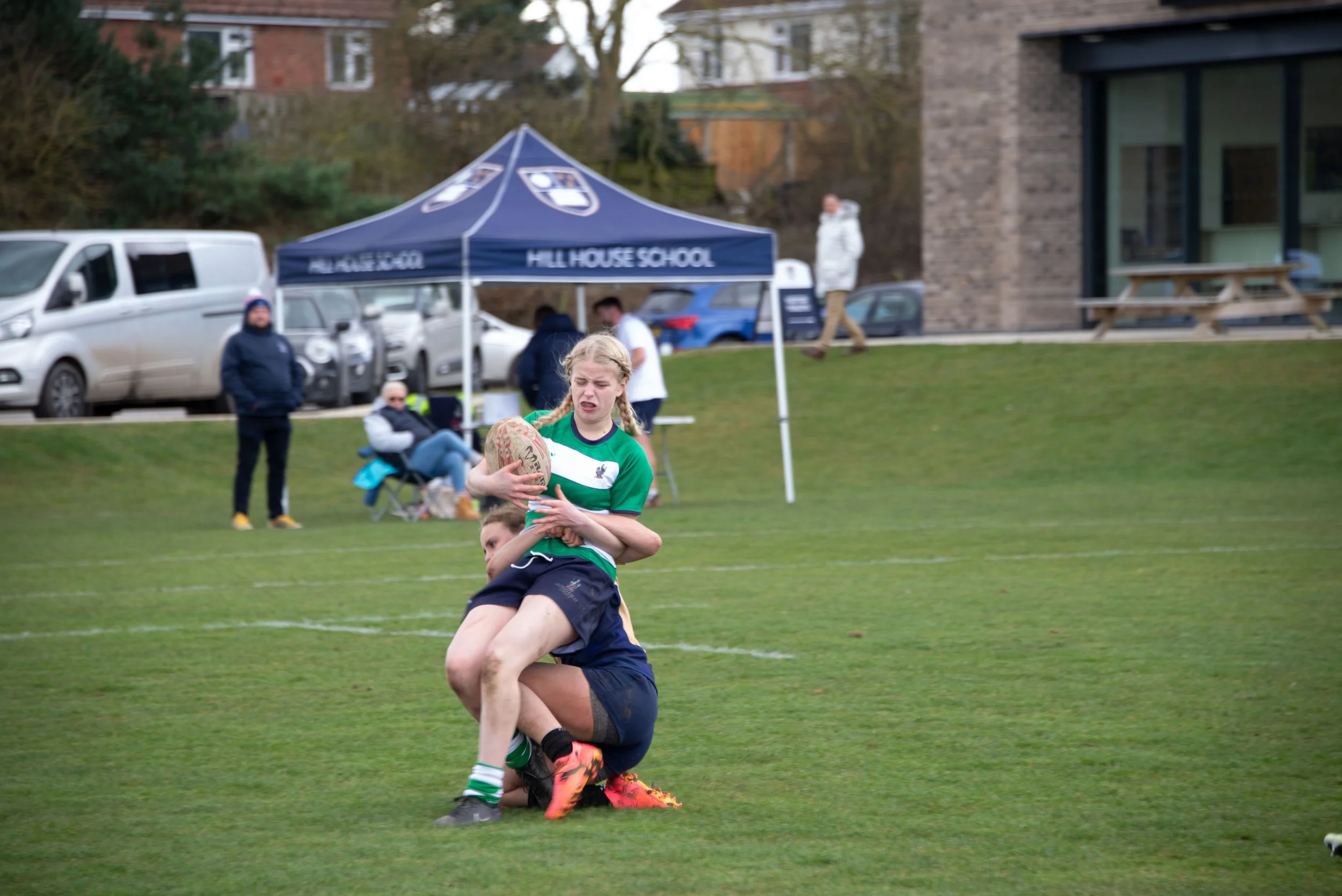 A young girl playing rugby tackle on a grassy field during a match, with a blue tent labeled 'Hill House School' and parked cars in the background.