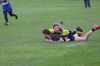 Two young girls playing rugby on a grassy field, with one girl tackling the other who is holding the ball.