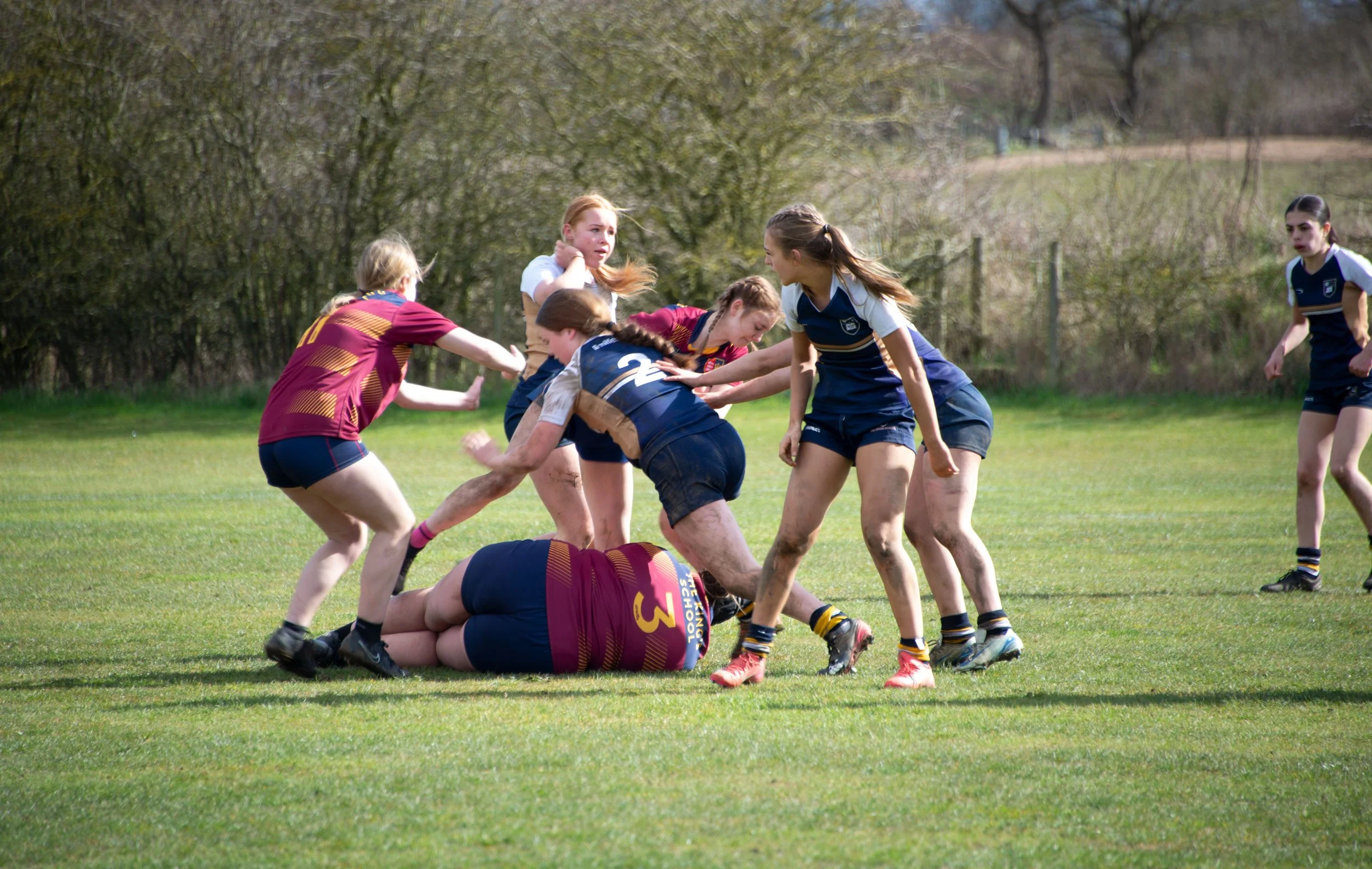 A women's rugby match with players fighting for the ball on a grassy field, some players are on the ground while others are reaching and pushing.