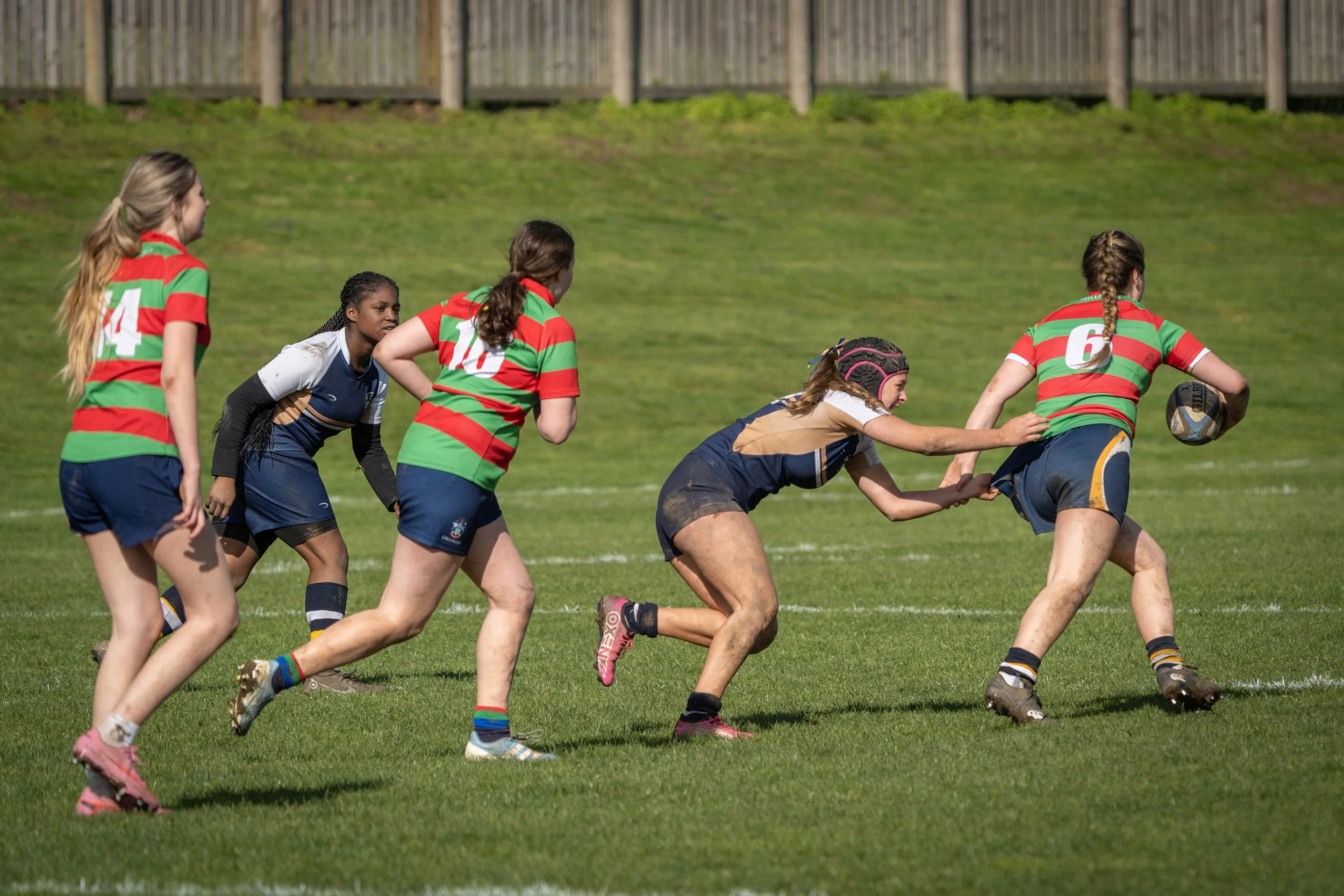Women playing rugby on a grassy field, with some wearing helmets and colorful striped uniforms.
