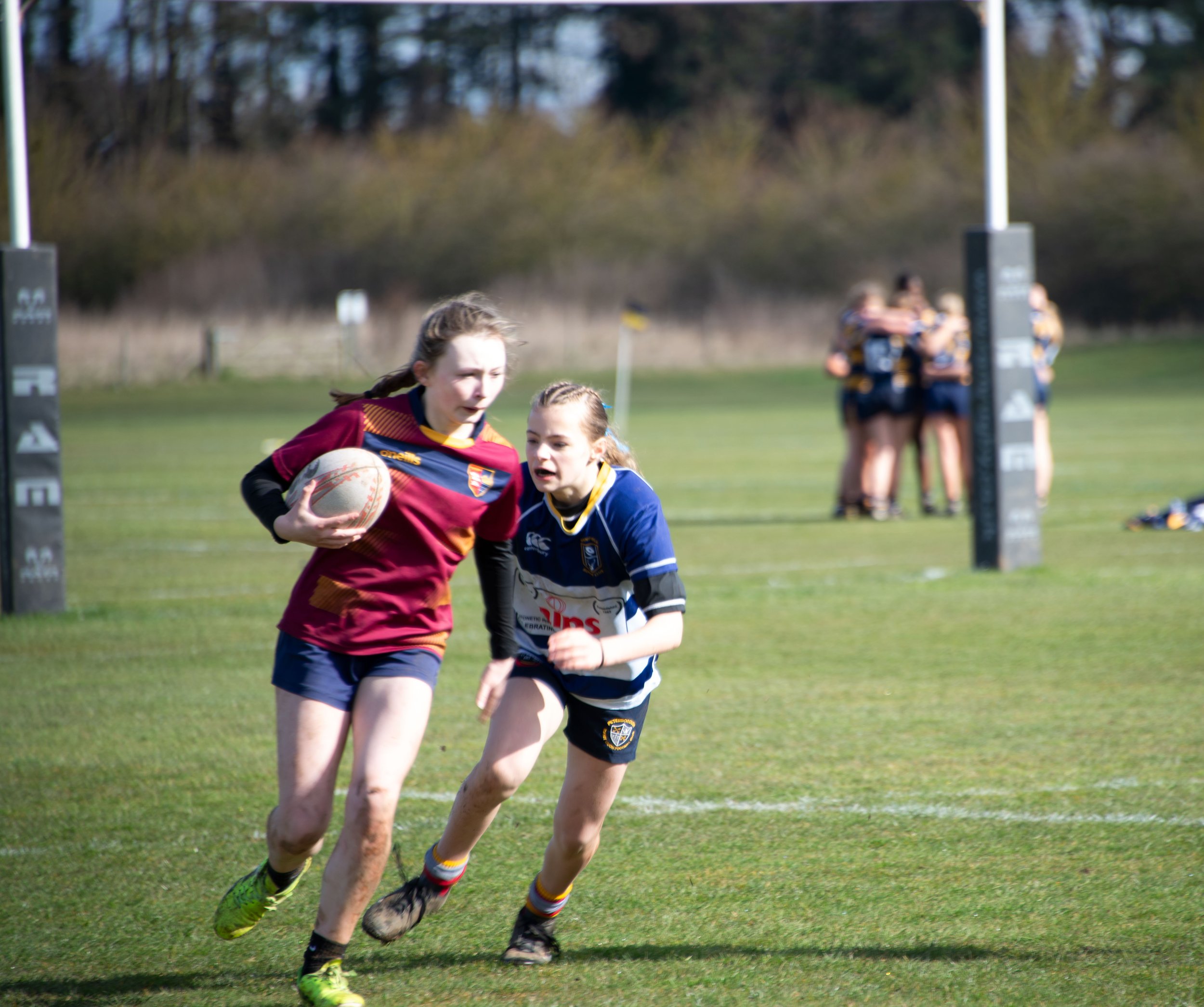 Young girls playing rugby on a grassy field, with a goal post in the background and a group of people watching.