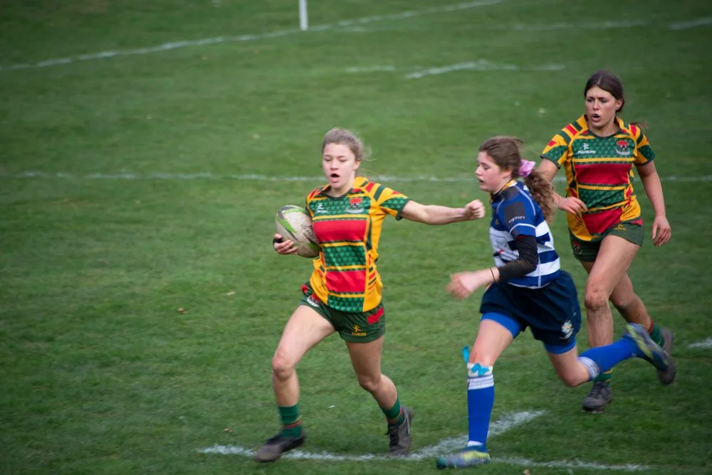 Three young girls playing rugby on a grassy field, one girl in a colorful yellow, red, and green jersey holding a rugby ball while being chased by two girls in blue and black jerseys.