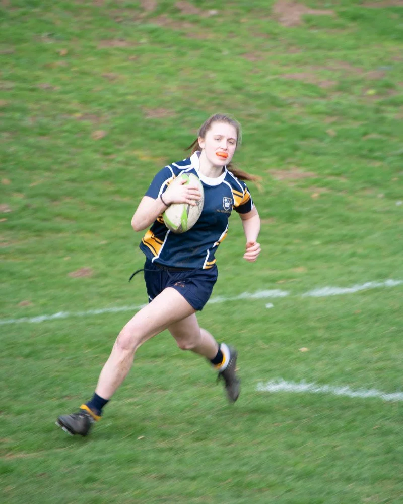 Young girl running on a grassy field while holding a rugby ball, wearing a blue sports uniform, with a determined expression.