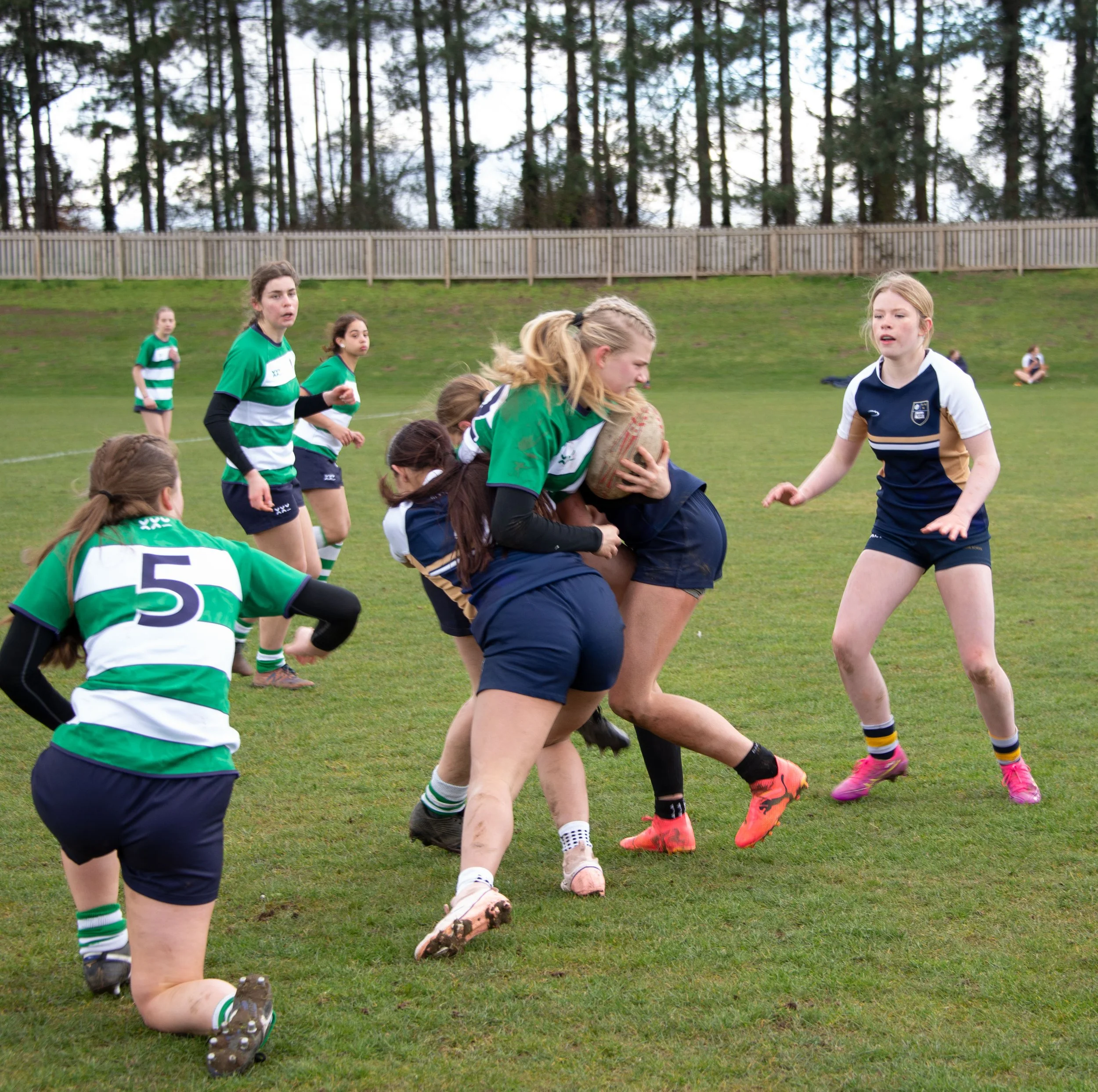 Group of young women playing rugby on a grassy field, with trees and a wooden fence in the background.
