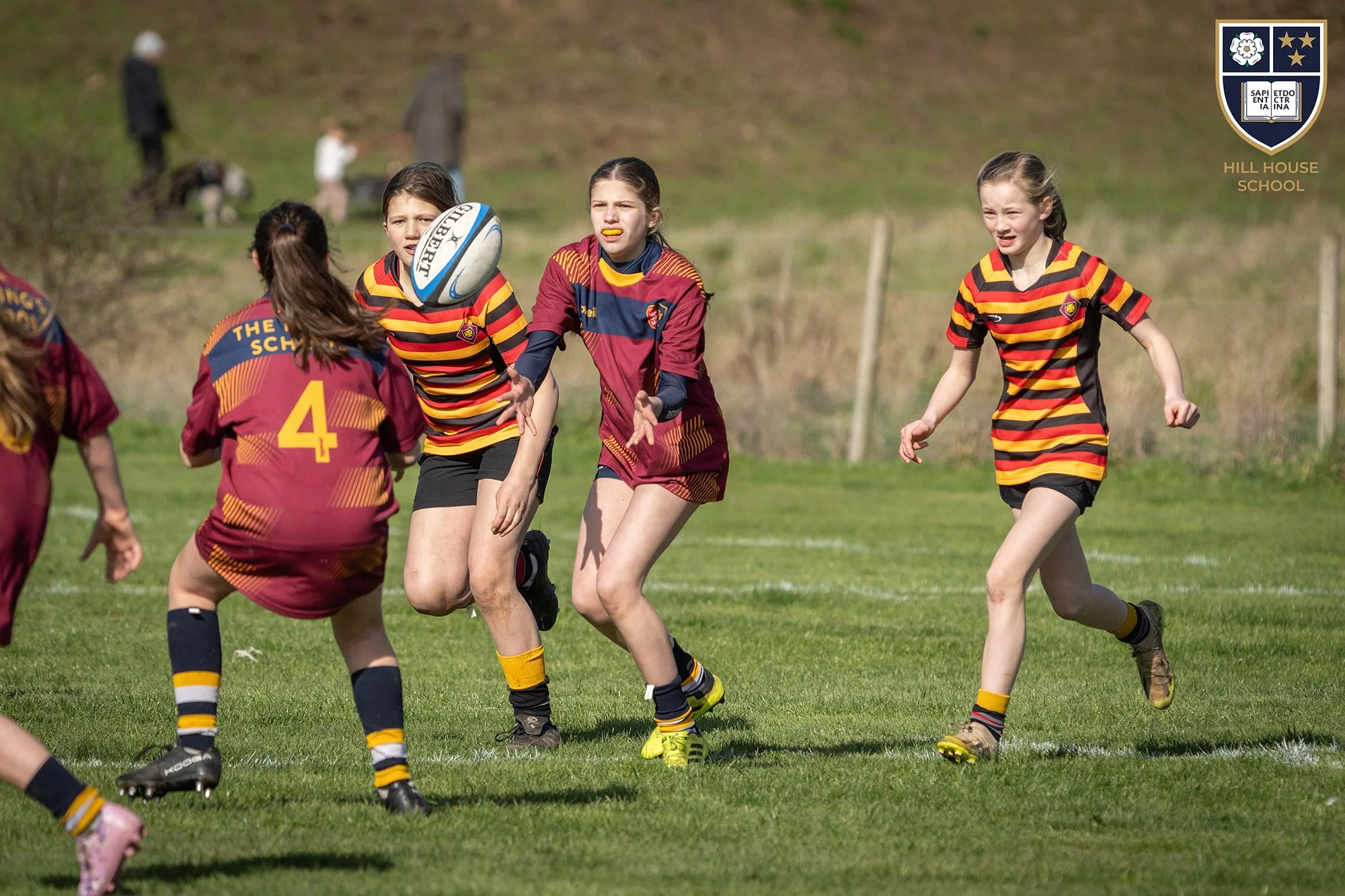 Young girls playing rugby on a grassy field, wearing maroon and yellow uniforms with the Hill House School crest, with a rugby ball in mid-air, during a sunny day.
