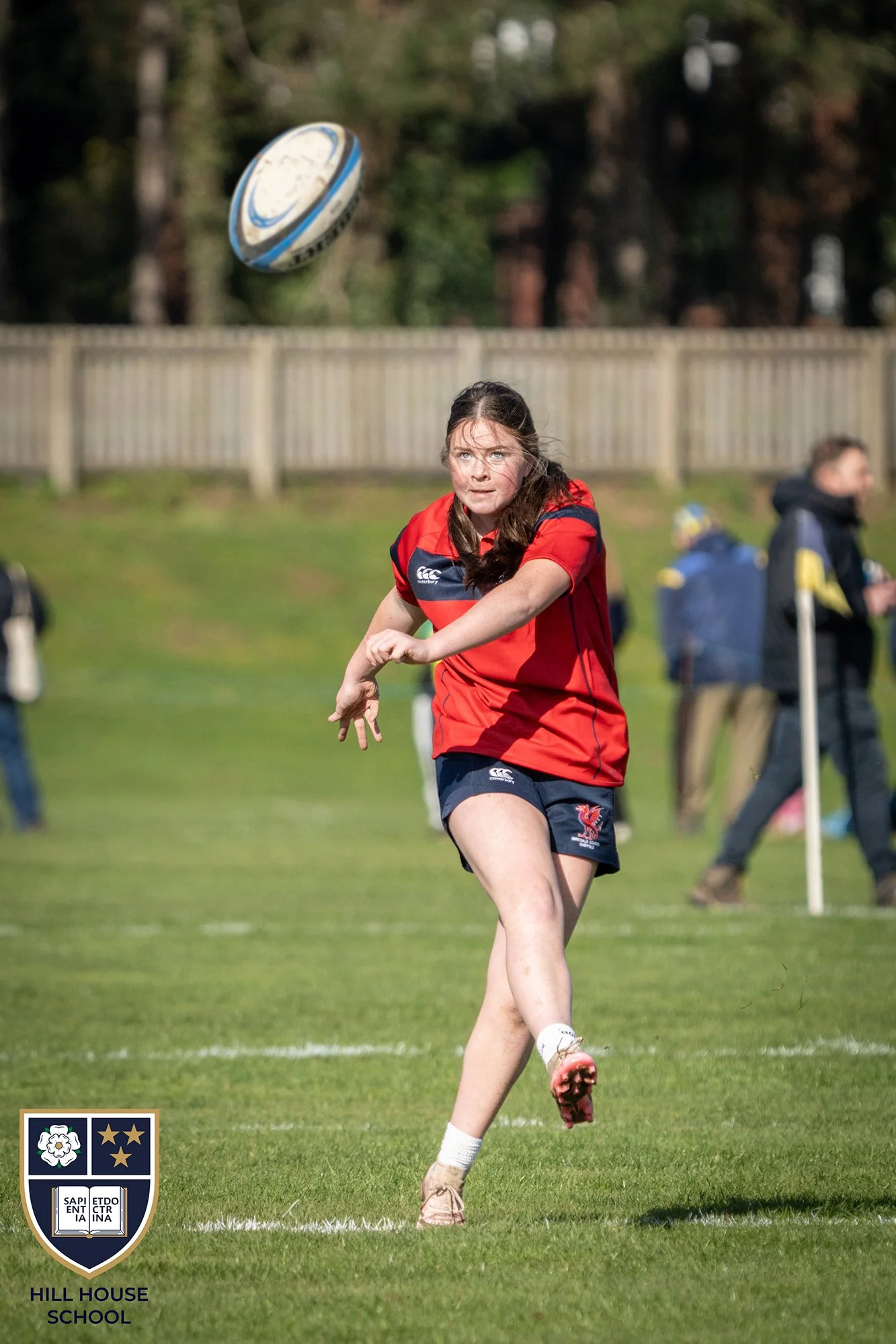A female rugby player in red and blue uniform running on a grass field during a game, with a rugby ball in mid-air, and a school crest with the words 'Hill House School' in the bottom left corner.
