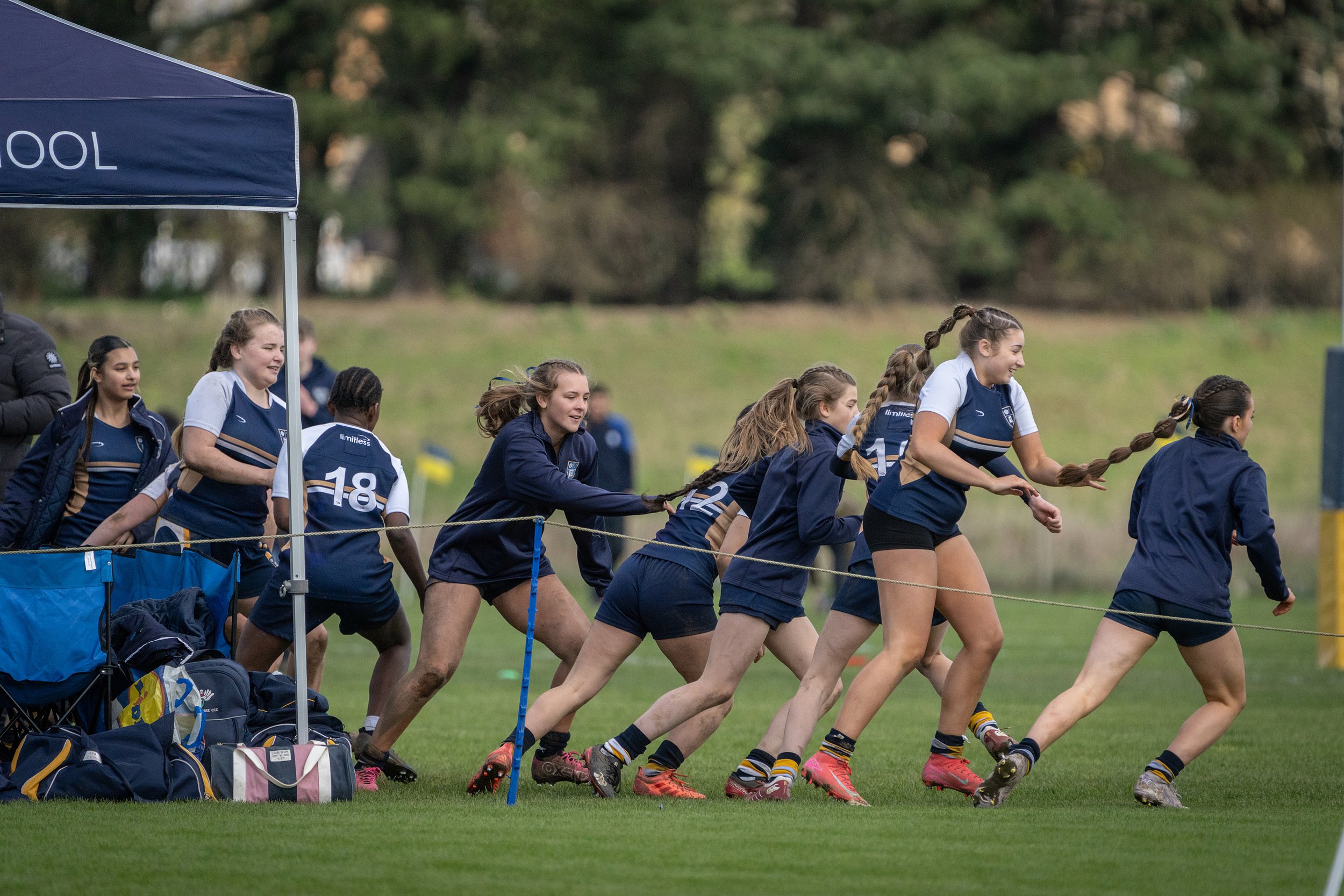 Girls in a rugby match running on a grassy field during daytime, with some sitting under a blue tent nearby.