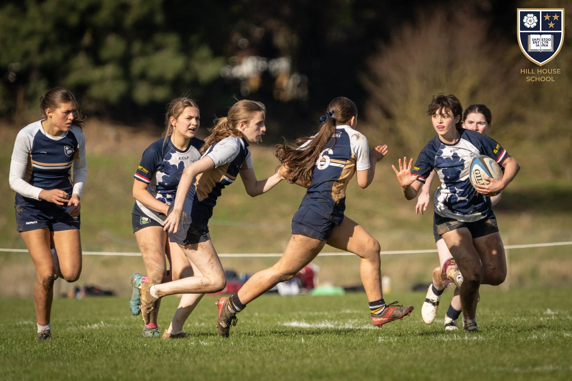 Girls playing rugby on a grassy field with muddy shoes and uniforms, with trees in the background and a school crest in the top right corner.