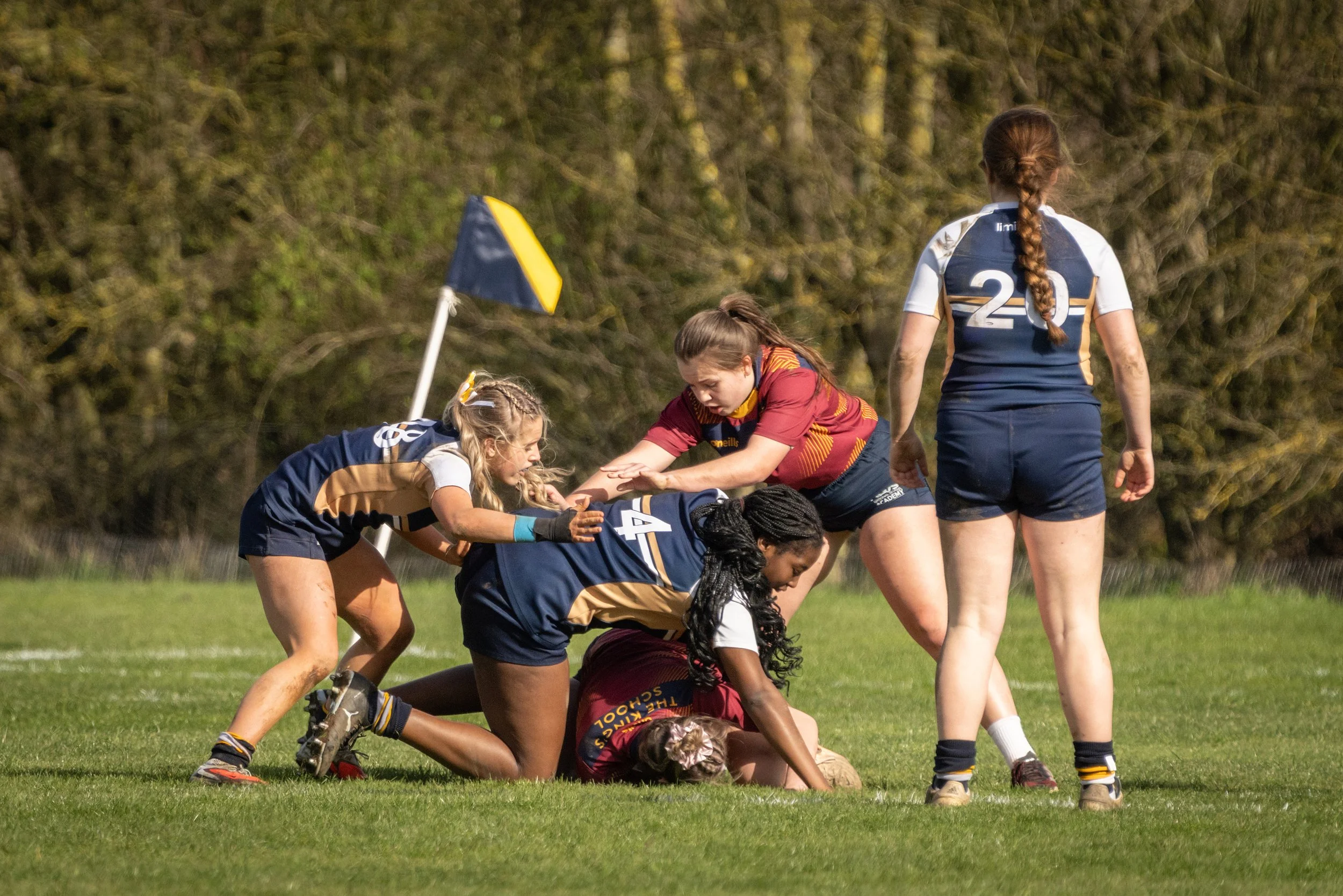 Women playing rugby on a grassy field with a yellow and blue corner flag, surrounded by trees.