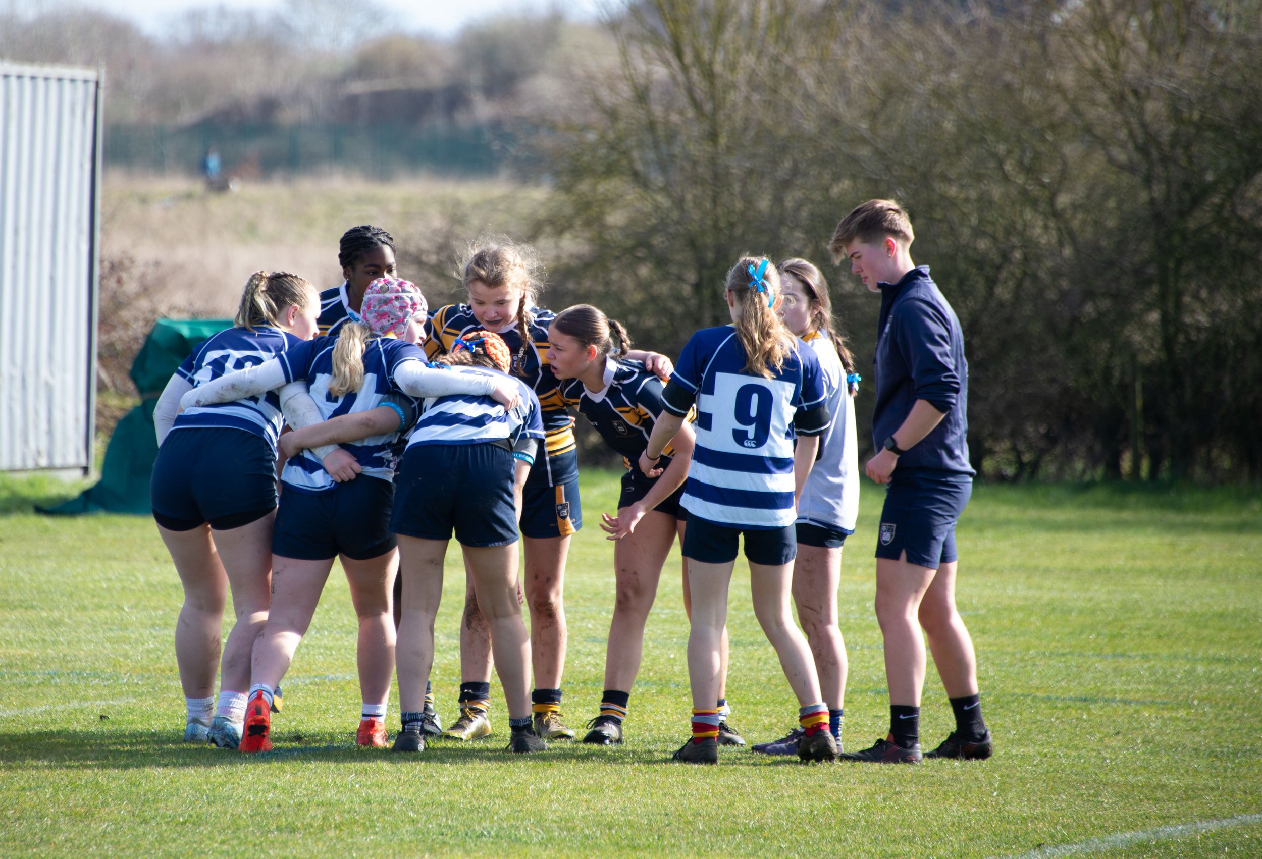 A girls' rugby team huddled together on a grassy field during a game, with their coach standing nearby.