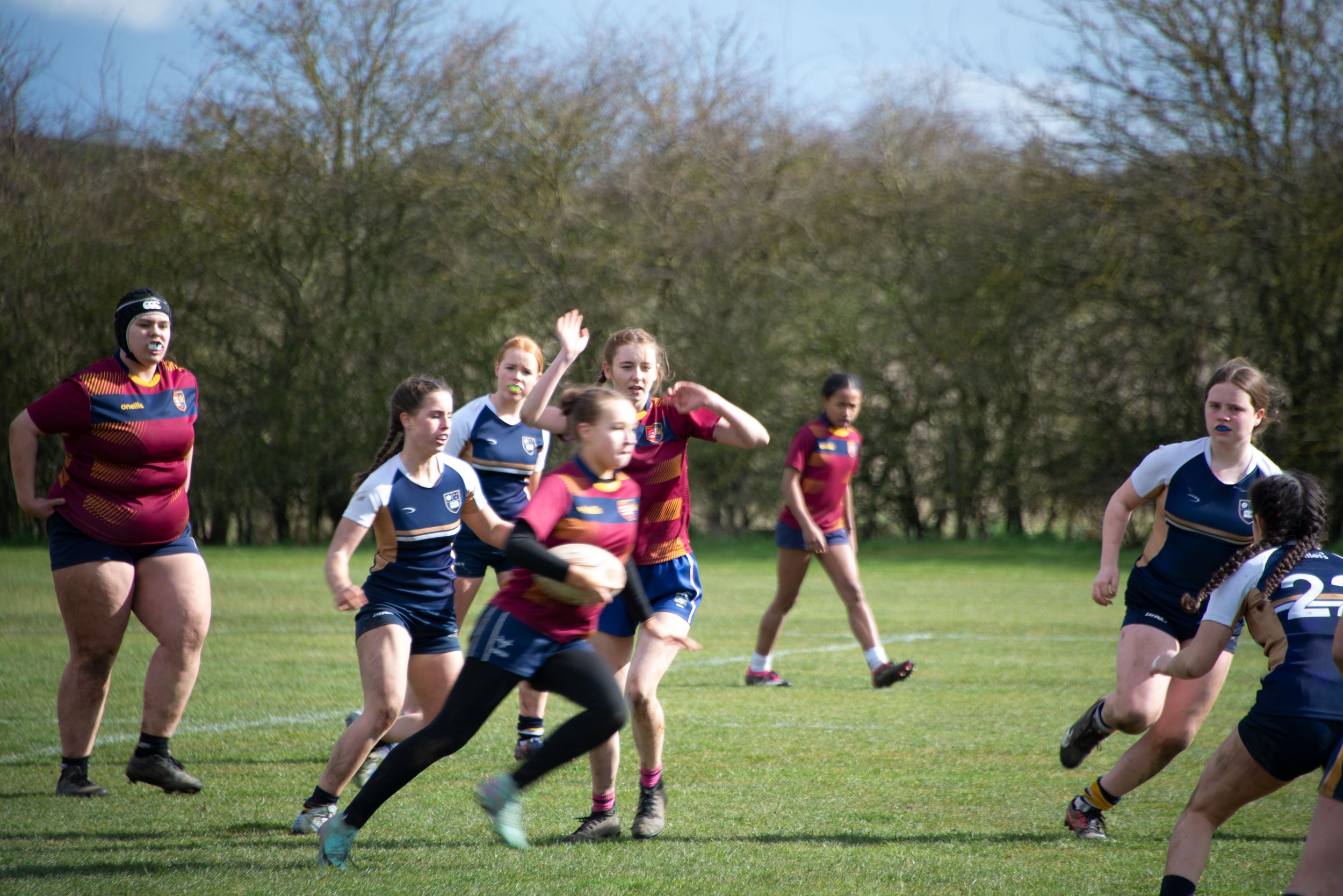 Girls playing rugby on a grassy field with trees in the background.