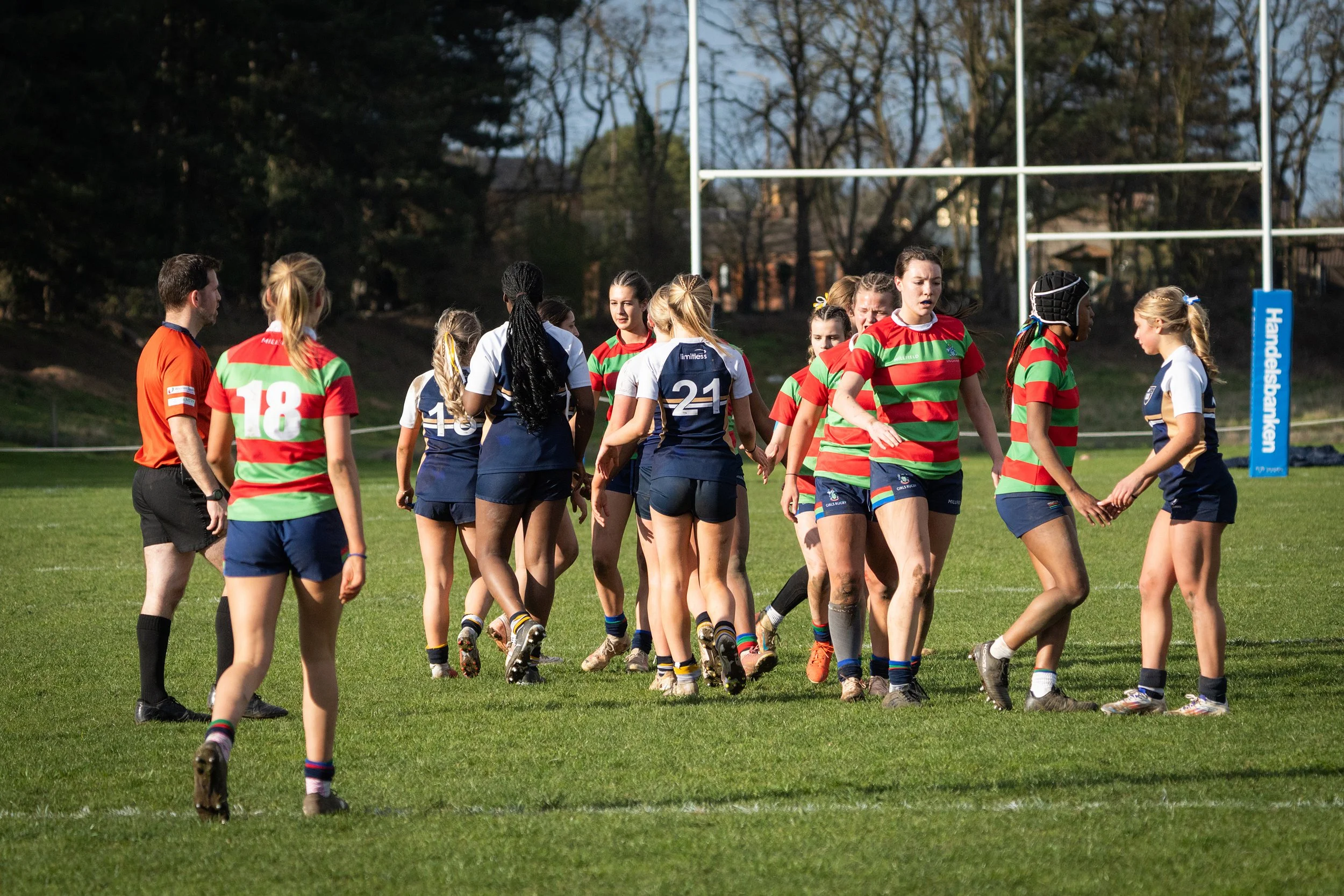 Rugby players on a field after a game, some wearing colorful jerseys, with goalposts in the background.