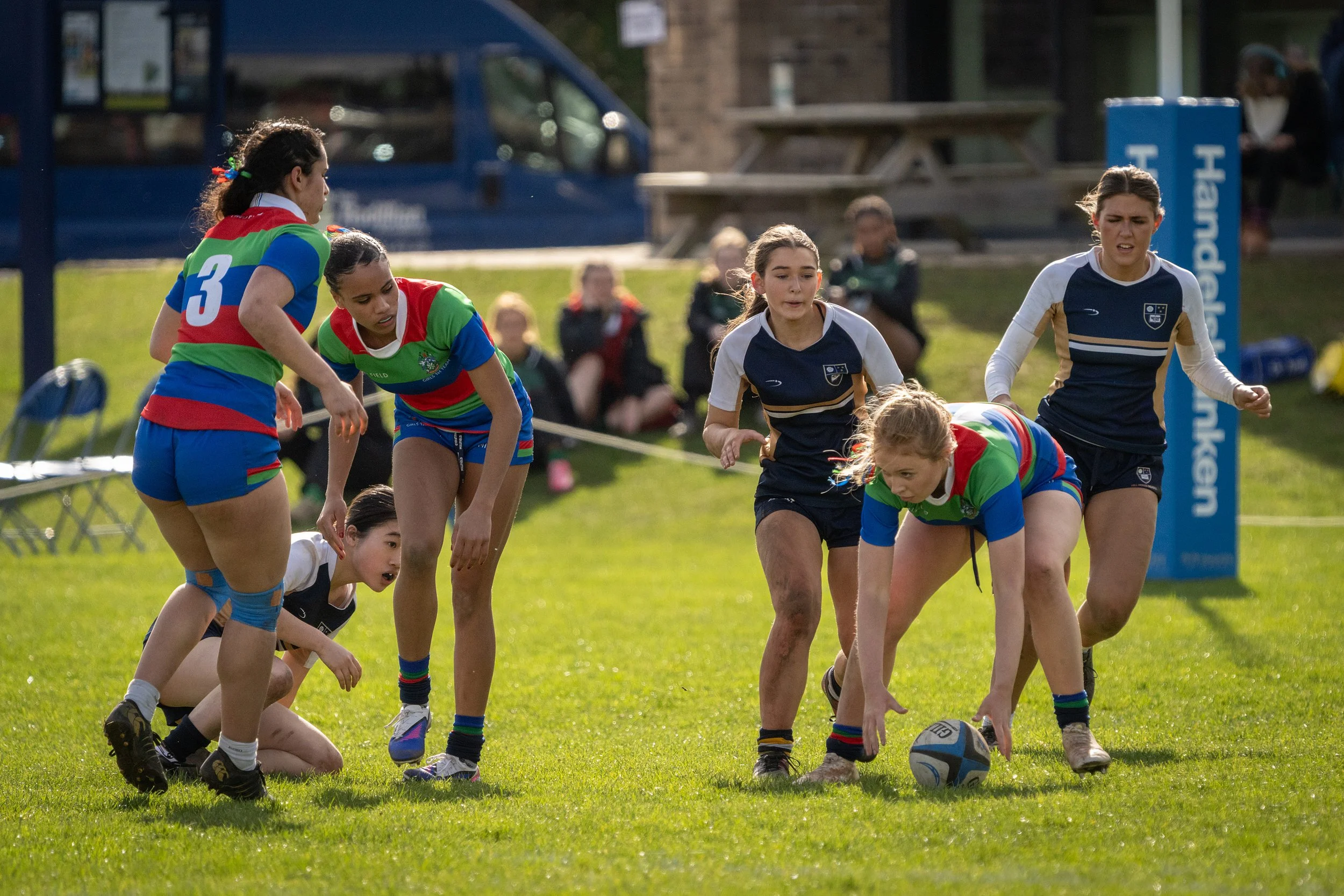 Women playing rugby on a grassy field, dressed in colorful jerseys, with spectators in the background.