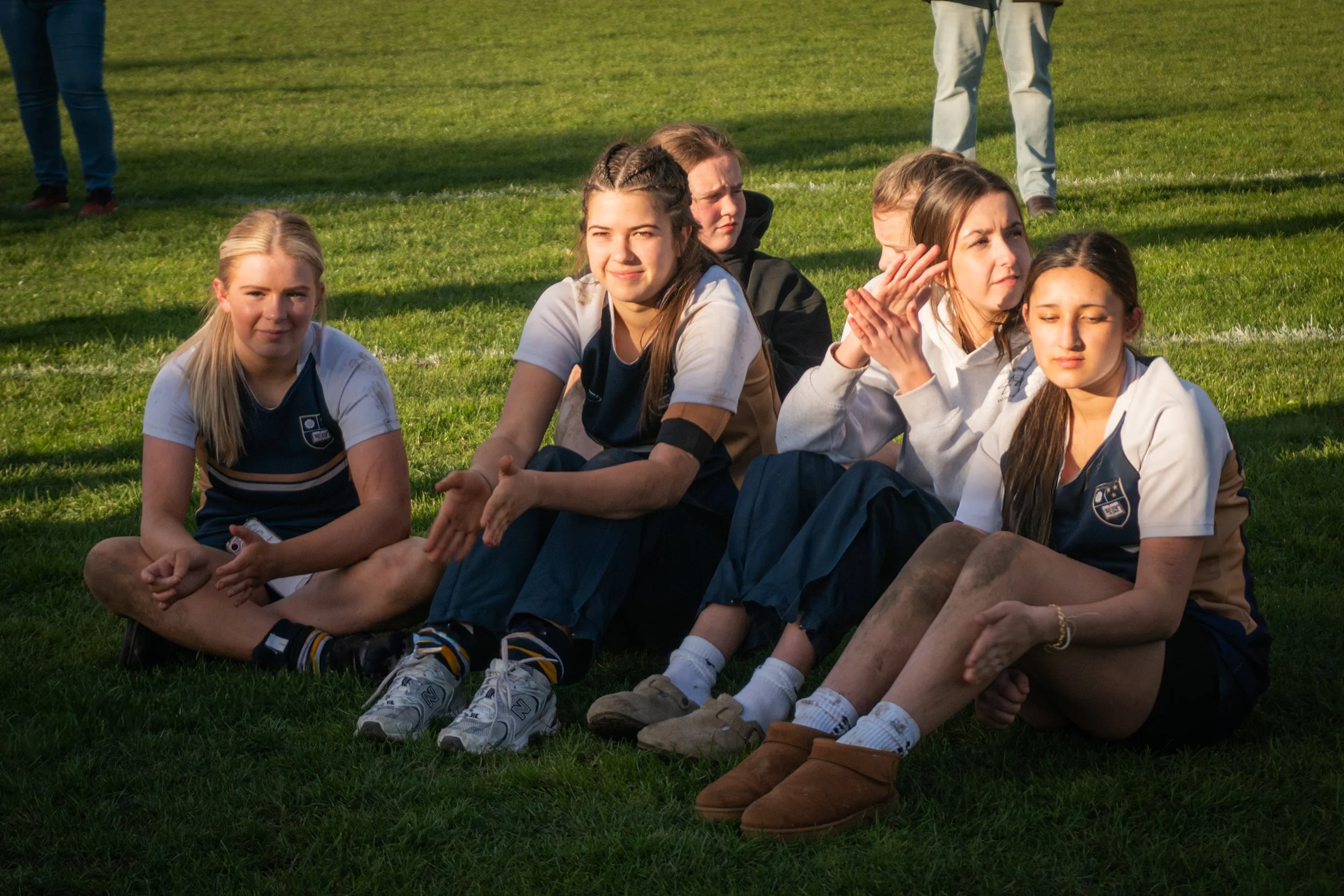Group of young women sitting on grass in outdoor setting, some clapping and looking in different directions, casual clothing, bright sunlight.