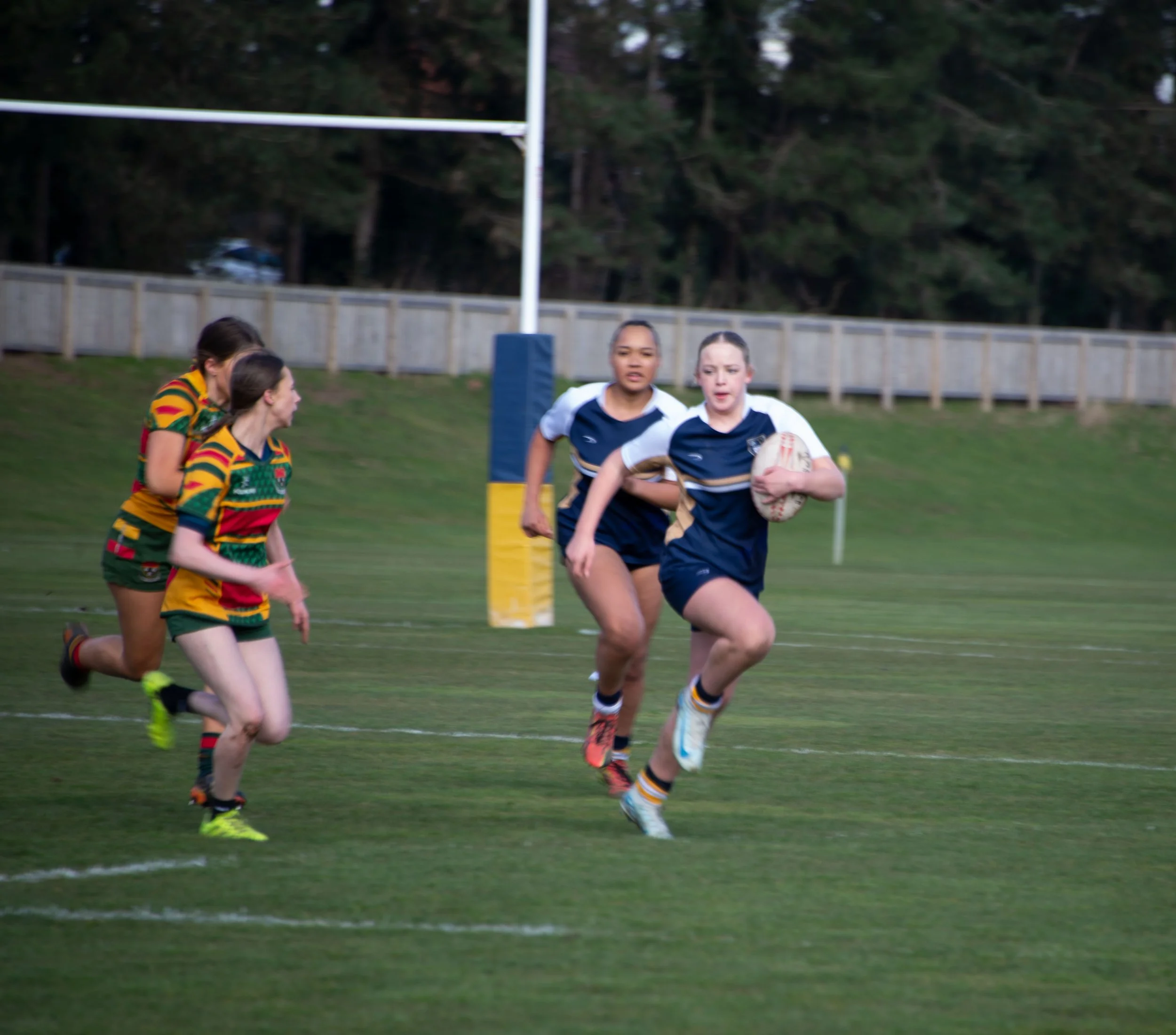 Rugby players running on a field with a rugby ball during a game, with a goal post and a wooden fence in the background.