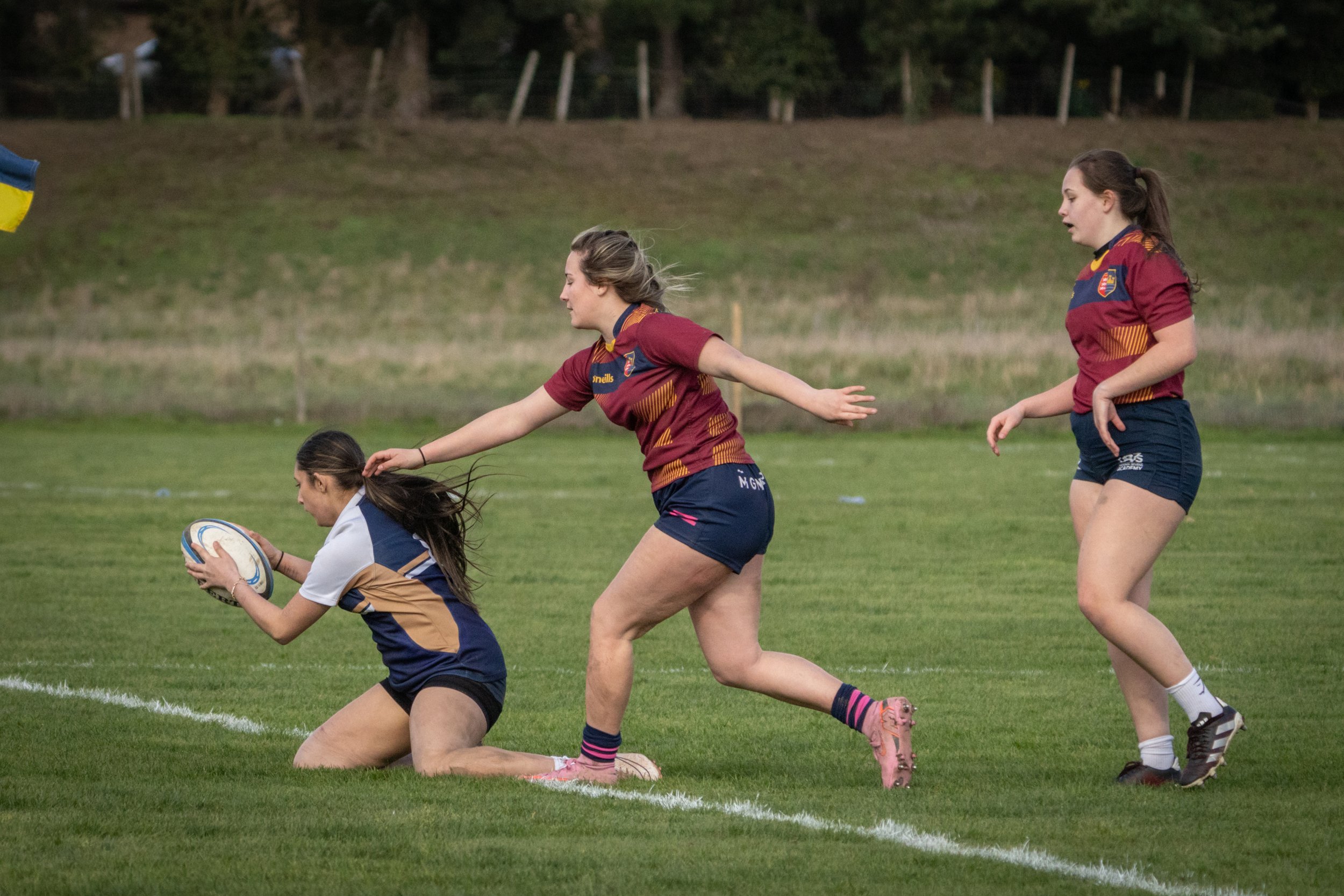 Women playing rugby on a grassy field, with one woman holding a rugby ball and the others approaching or defending.