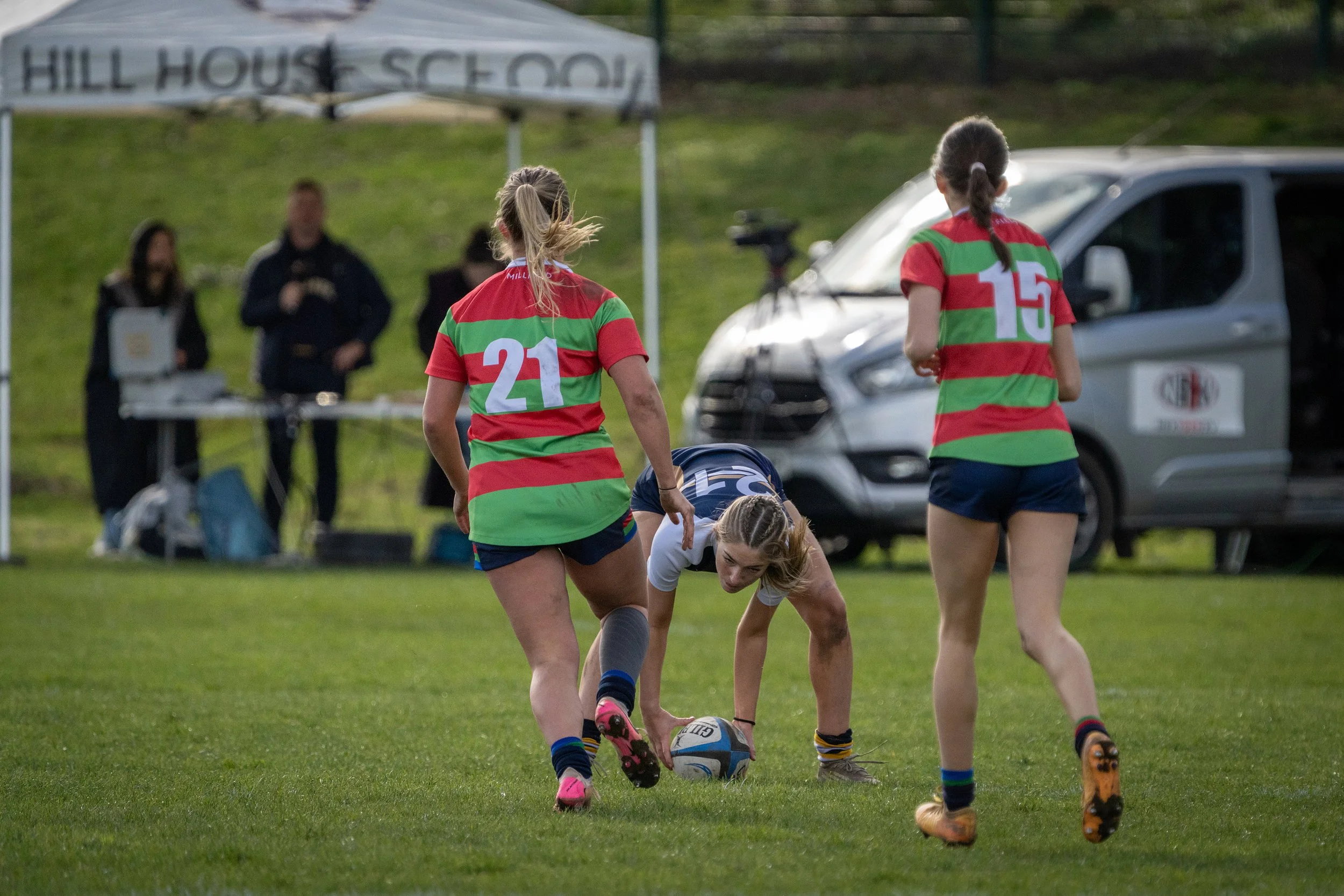 Two female rugby players wearing green and red striped jerseys and blue shorts are on the field, one is bending down and the other stands near her, both are focused on the rugby ball on the grass.