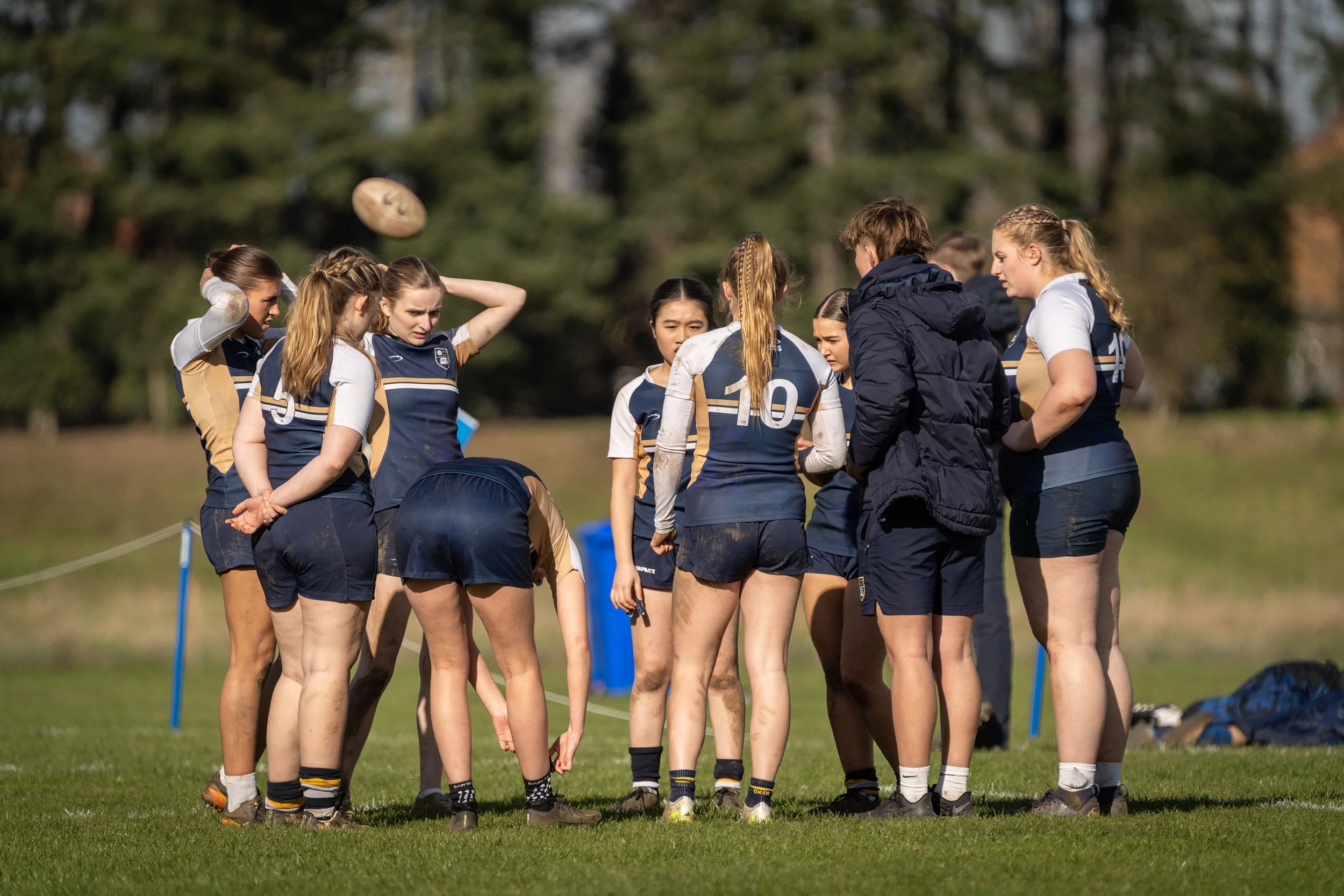 A group of female rugby players listening to their coach during a match or practice on a grassy field, wearing matching jerseys in navy blue and tan, with some players having muddy uniforms.