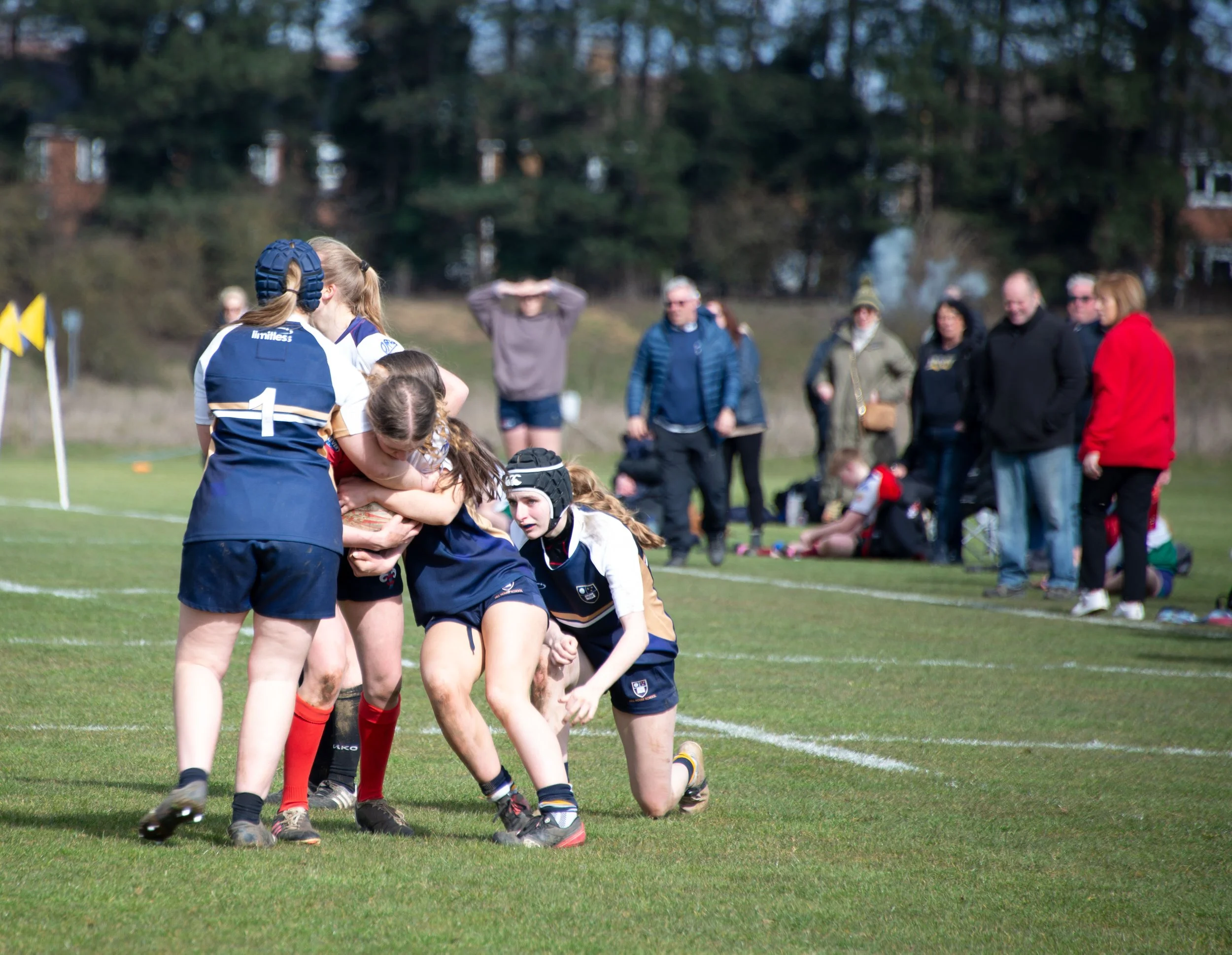 A Girls rugby match on a grassy field showing a pile-up of players with referees and spectators in the background.