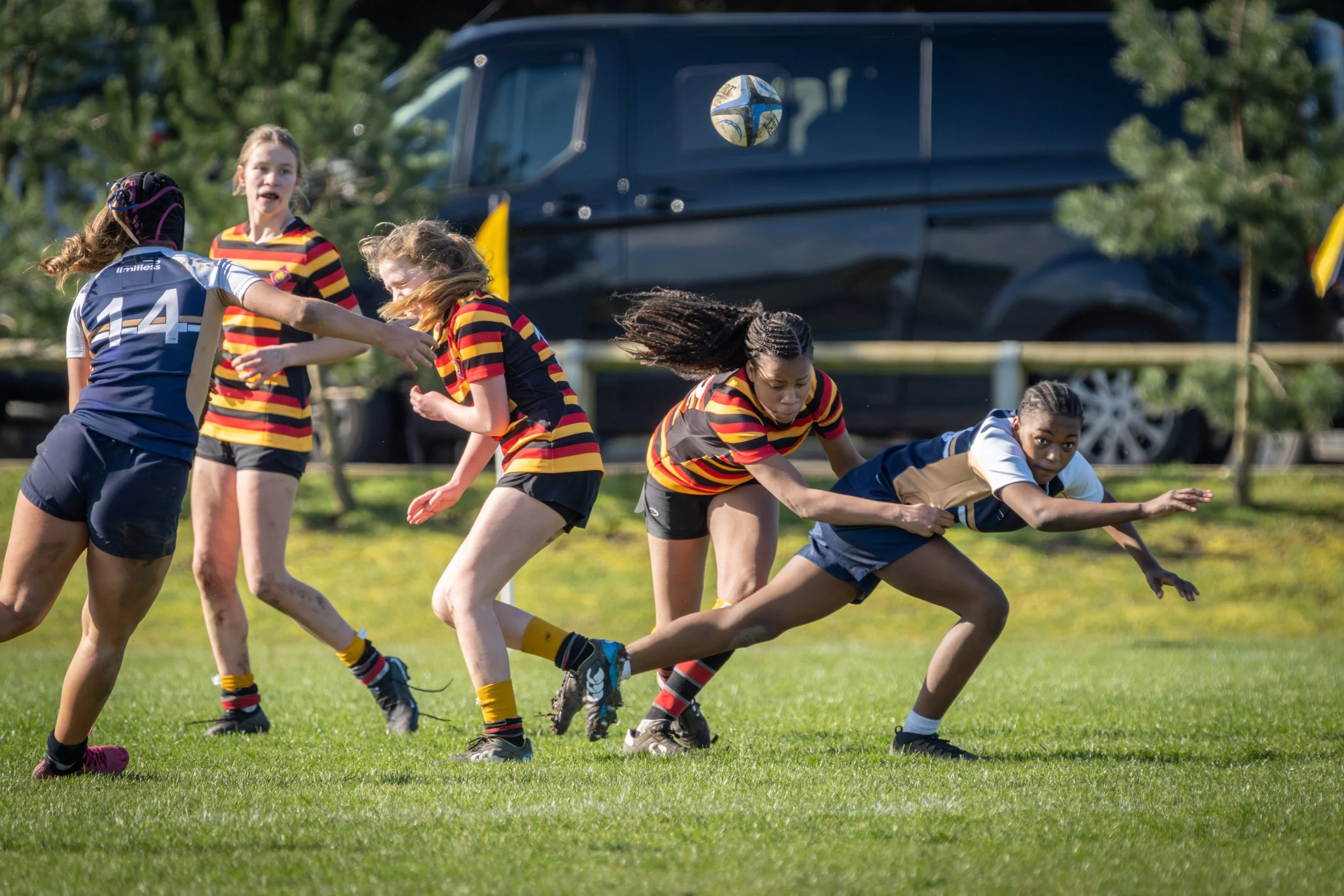 Children playing rugby on a grassy field with a football in the air, trees, and a parked black vehicle in the background.