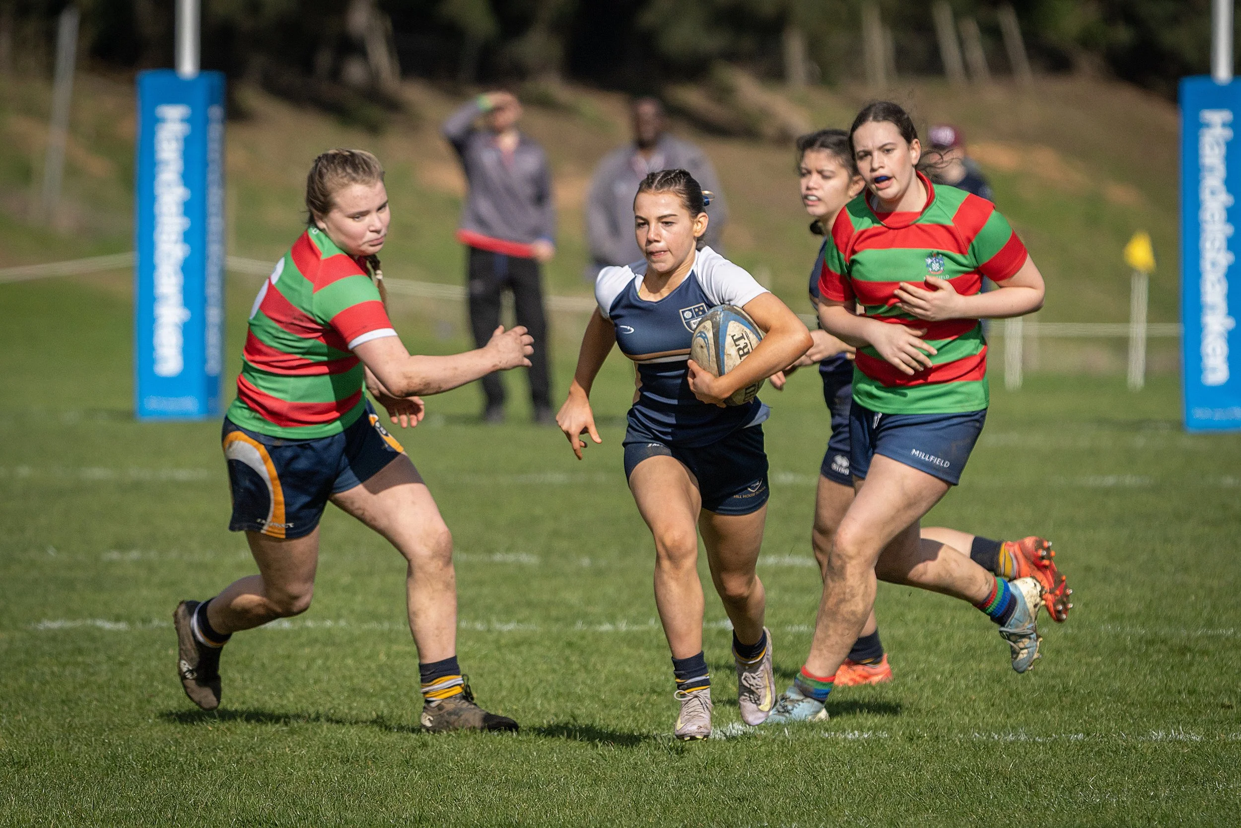 Women playing rugby on a grass field, with some women running and holding a rugby ball, wearing athletic uniforms and colorful socks, with spectators in the background.