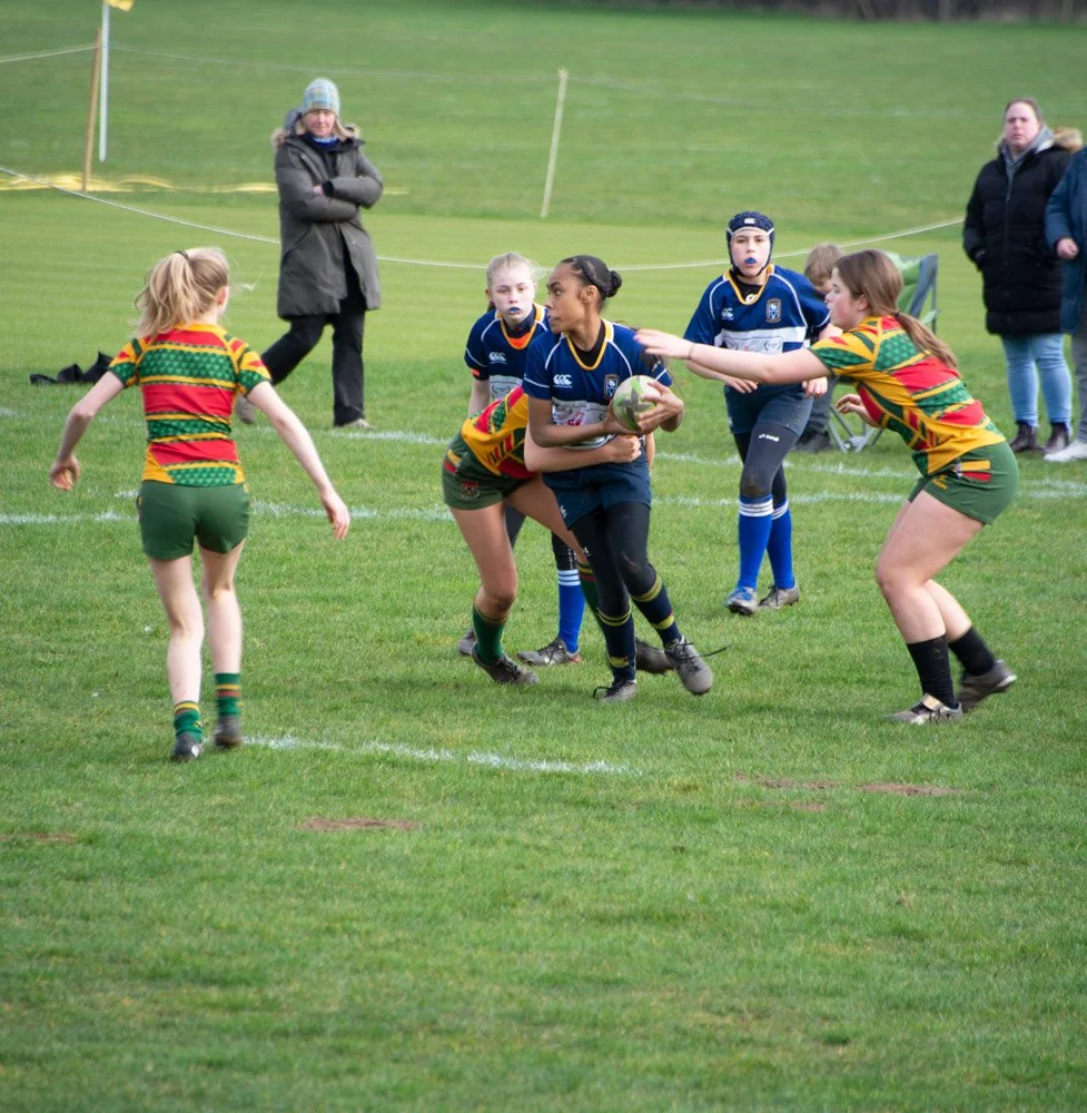 Girls rugby match on a grassy field, players wearing colorful jerseys, some in blue and others in red, with spectators watching in the background.