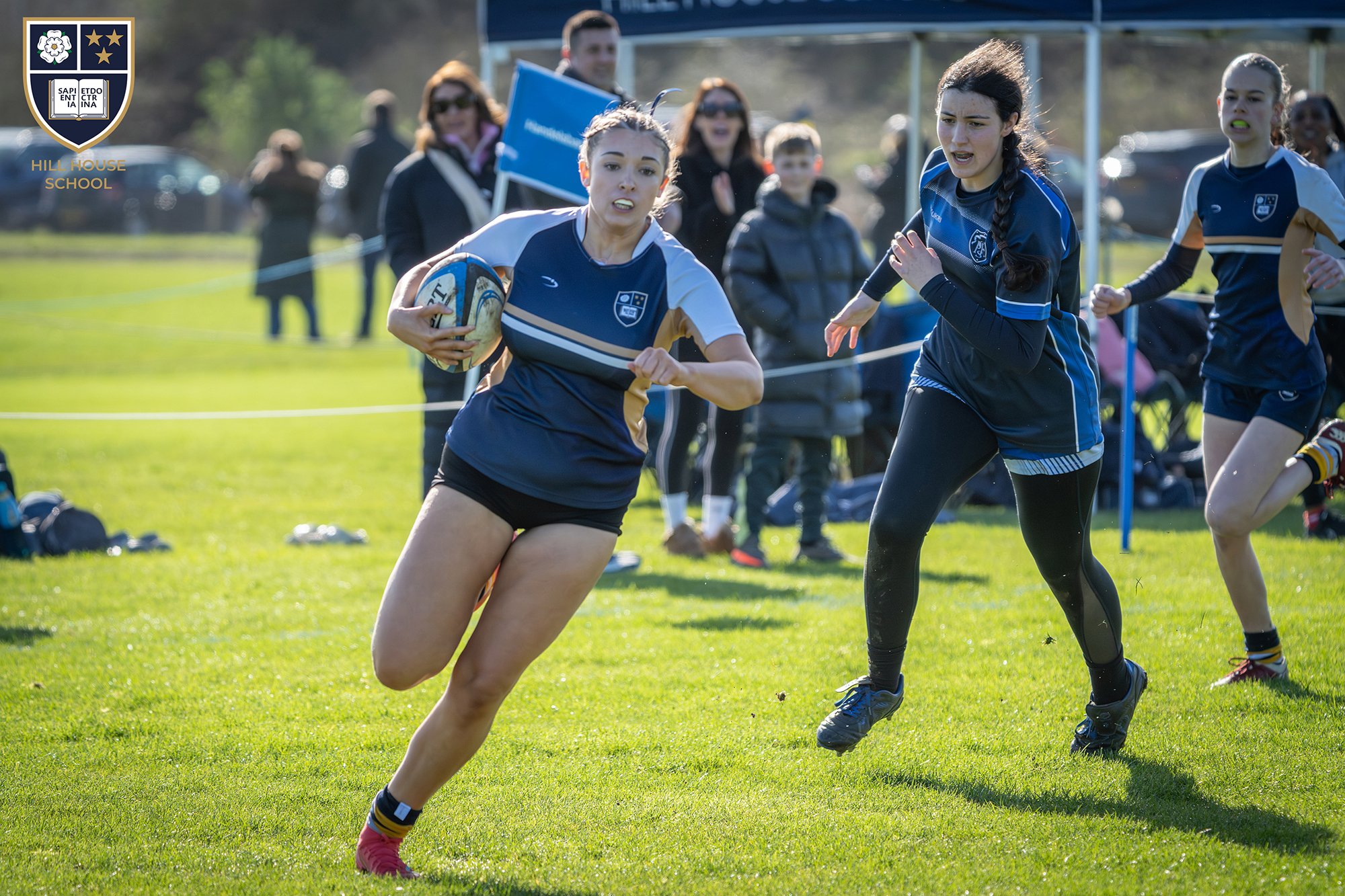 A girl wearing a blue and white sports jersey is running with a rugby ball during a game on a grass field. Other players and spectators are visible in the background.