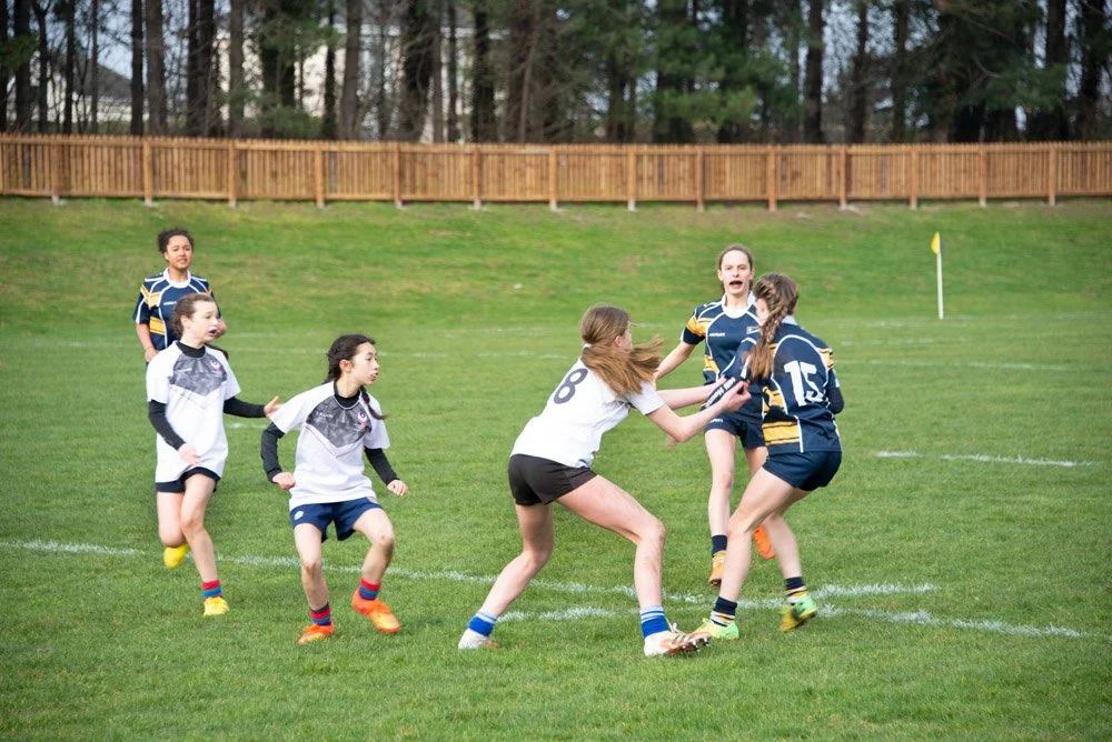Girls playing rugby on a grassy field with some trees and a wooden fence in the background.