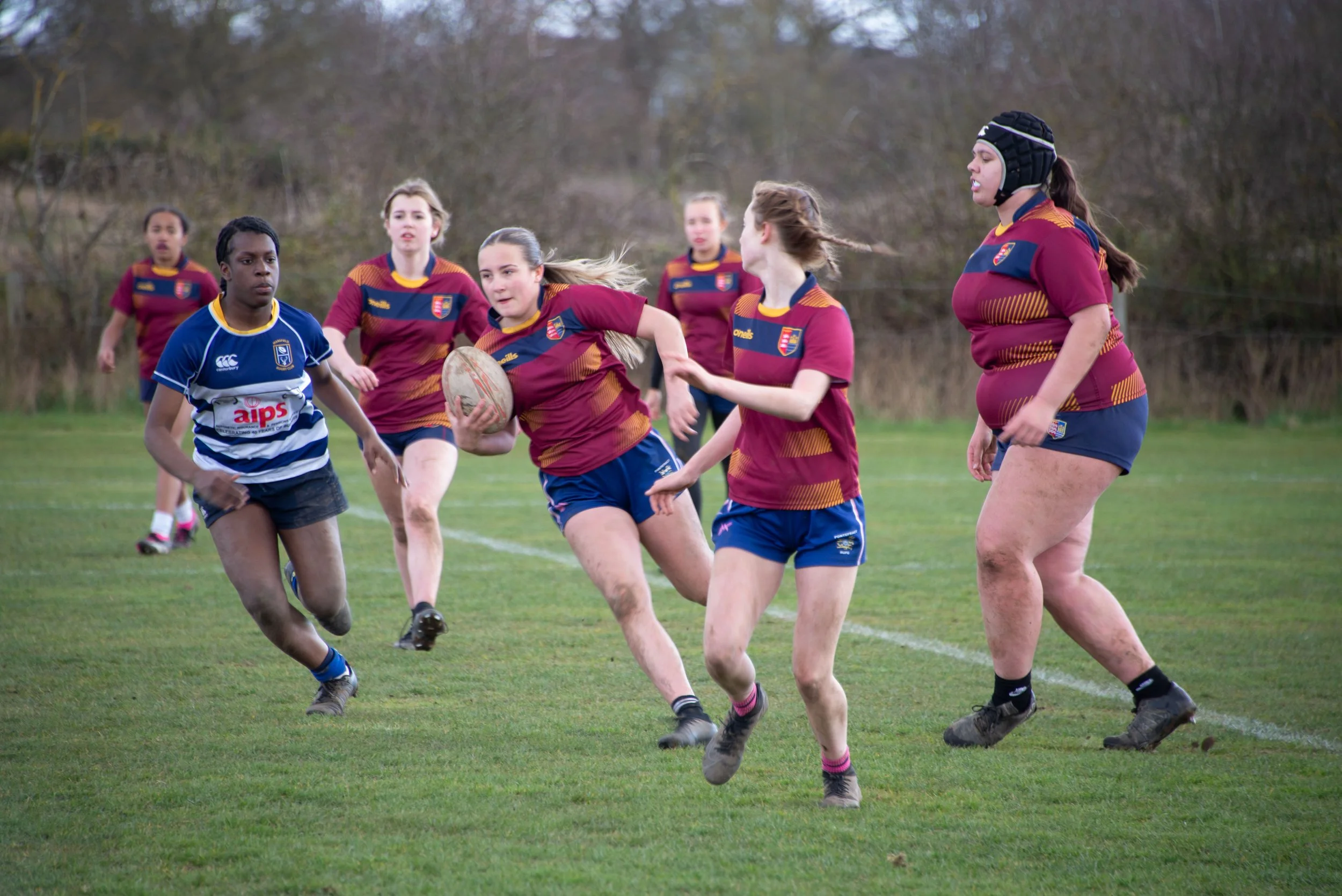 Women playing rugby on a grassy field, with some women running and others preparing to catch or tackle the ball, dressed in colorful uniforms.