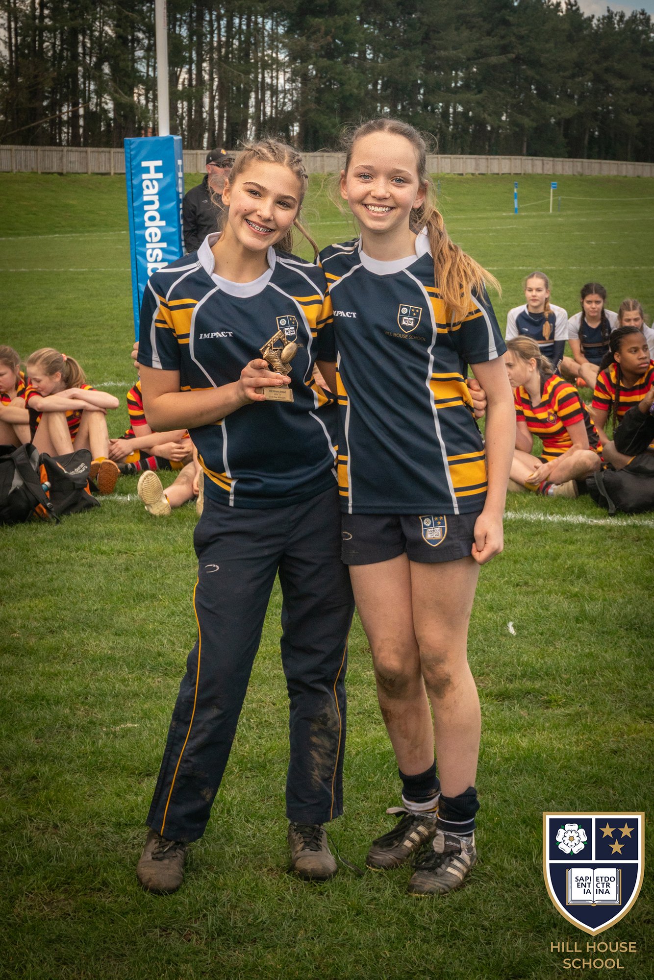 Two girls in sports uniforms standing on a grassy field, smiling, with one holding a trophy. Other children are sitting in the background, and a school crest with the text 'Hill House School' is in the lower right corner of the image.