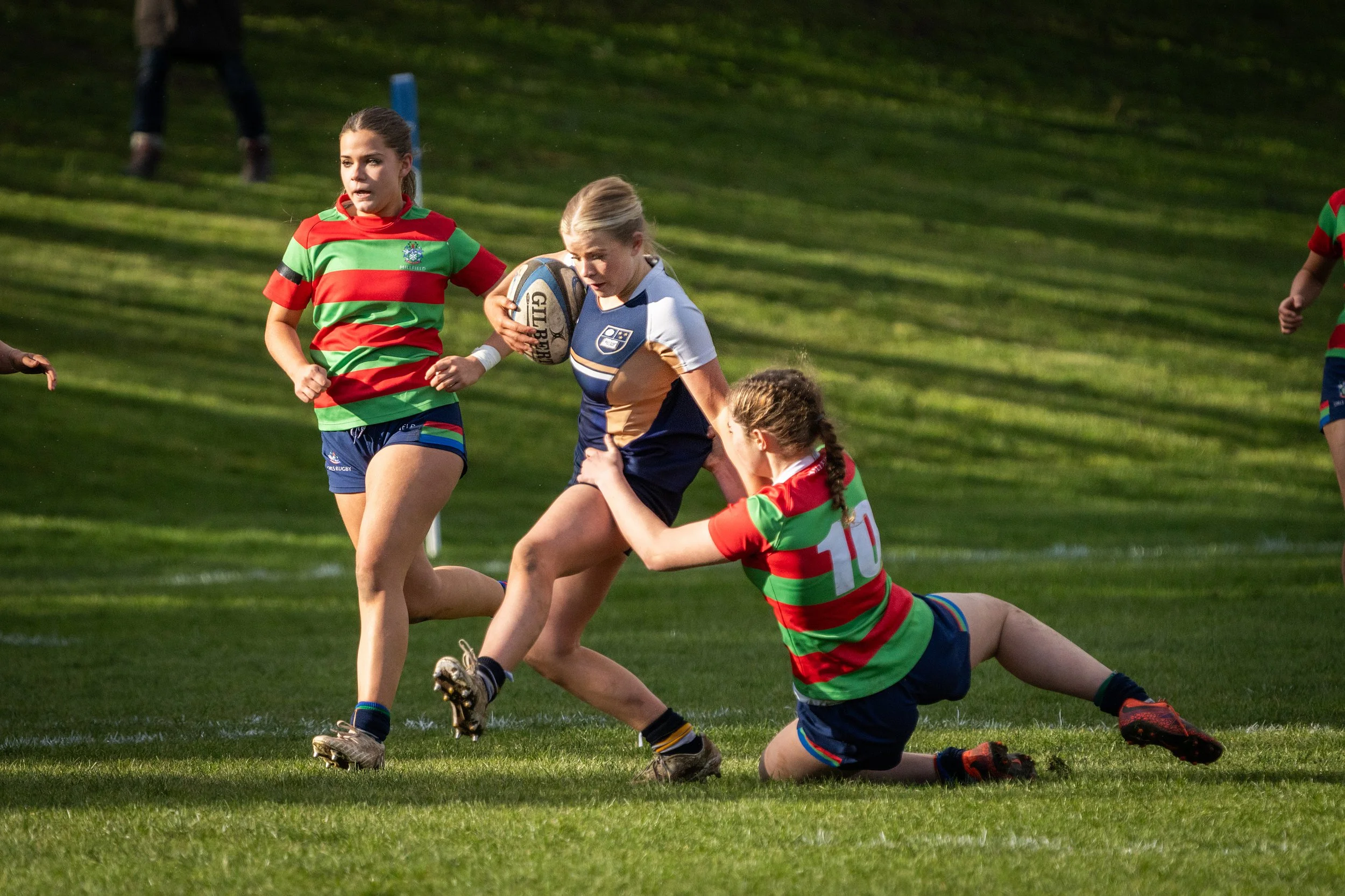 Women playing rugby on a grassy field, with one holding a rugby ball and others tackling or defending.