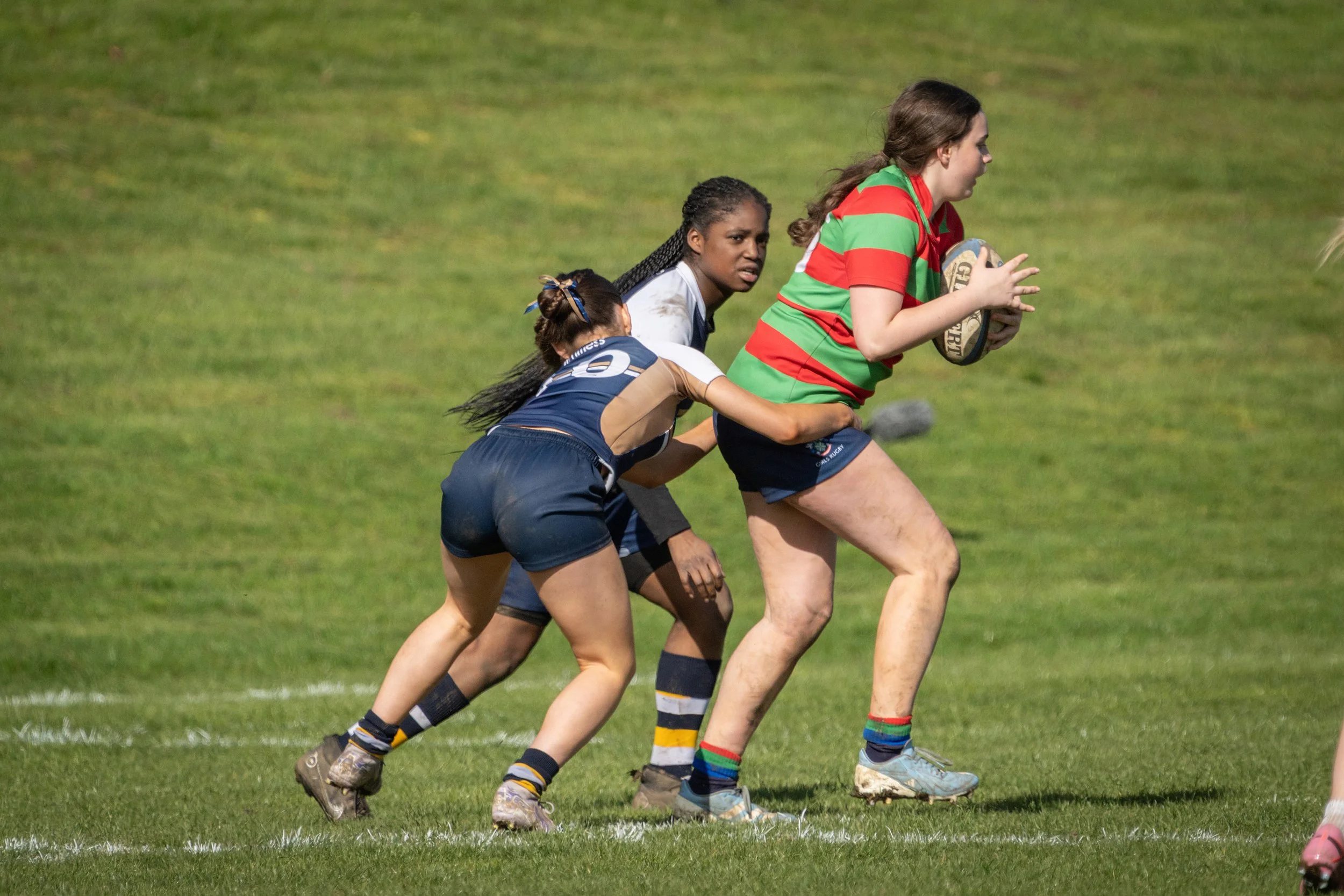 Women playing rugby on a grassy field, running with the ball, in sports uniforms.