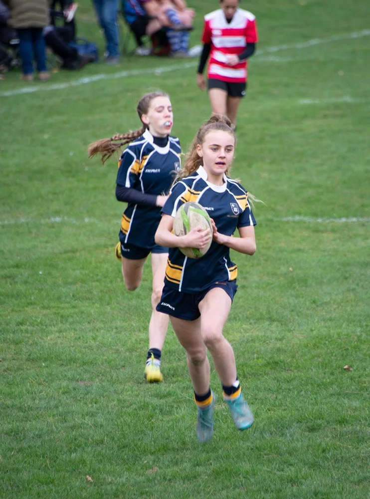 Young girls playing rugby on a grassy field with spectators in the background.