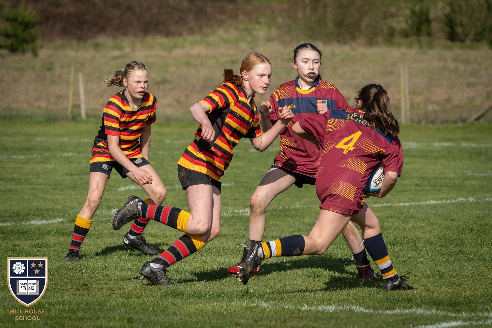 Girls playing rugby on a grassy field, wearing colorful striped uniforms, with one girl holding a rugby ball. There are trees and a hill in the background. The logo of Hill House School is visible in the bottom left corner.