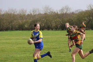 Children playing rugby on a grassy field with trees in the background.