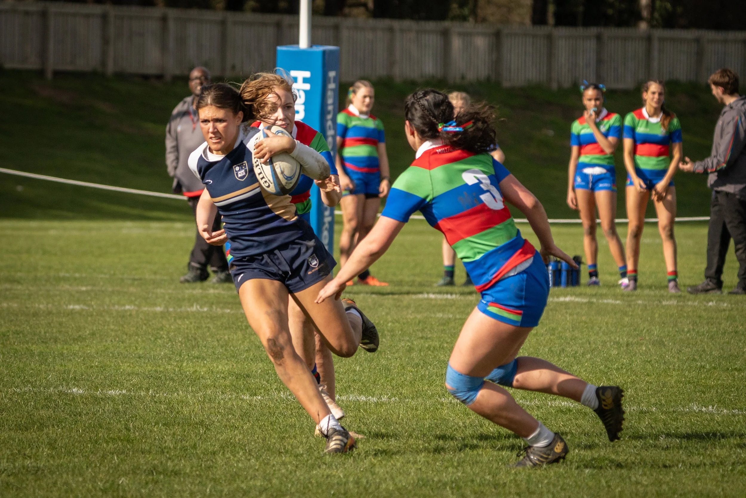 Women playing rugby on a grassy field, with one woman holding a rugby ball and others running around her. Spectators and team members are visible in the background, some in colorful uniforms.