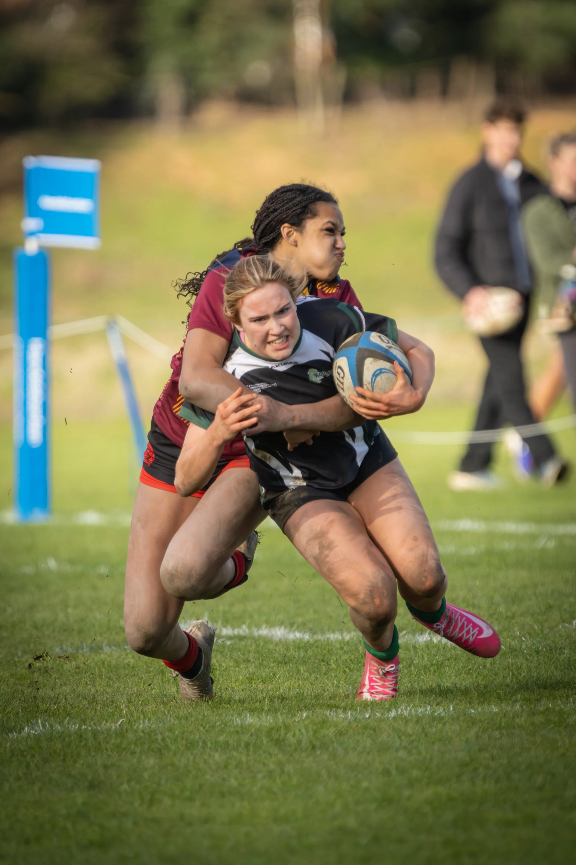 Two female rugby players, one in a maroon jersey and the other in a black and white jersey, fighting for possession of a rugby ball during a match on a grassy field. The player in black is holding the ball tightly while the player in maroon is trying to tackle her from behind. Blurred spectators can be seen in the background.