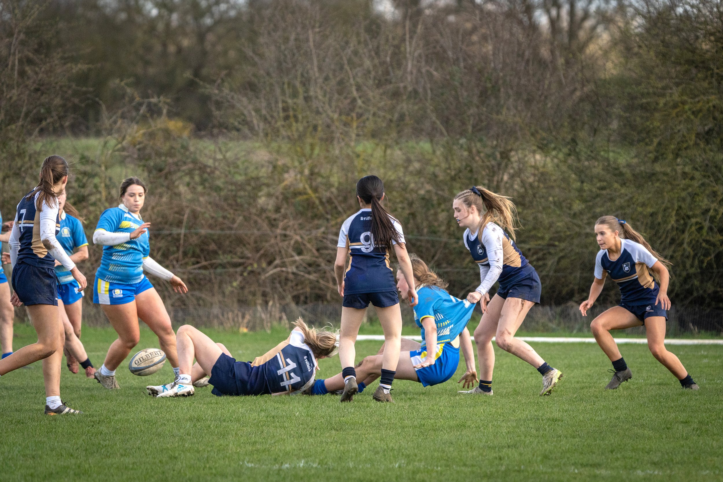 Women soccer players on a green field during a match, some are standing, others are on the ground, and a referee or coach is visible in the background.