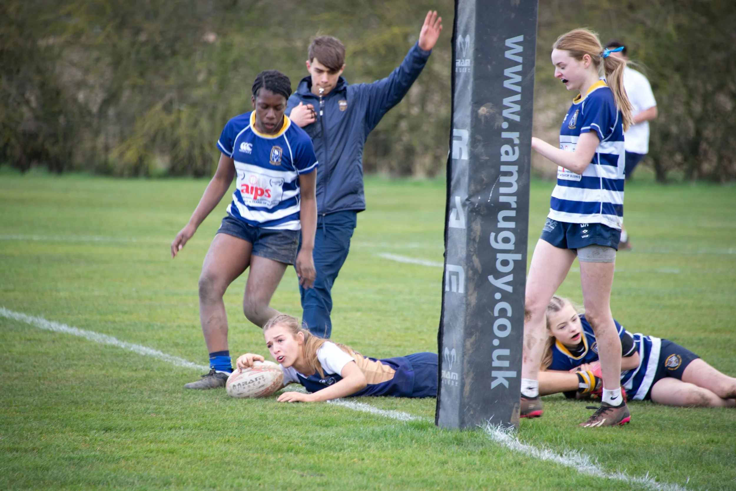 A group of young girls playing rugby on a grassy field, with some players lying on the ground and a referee or coach nearby.