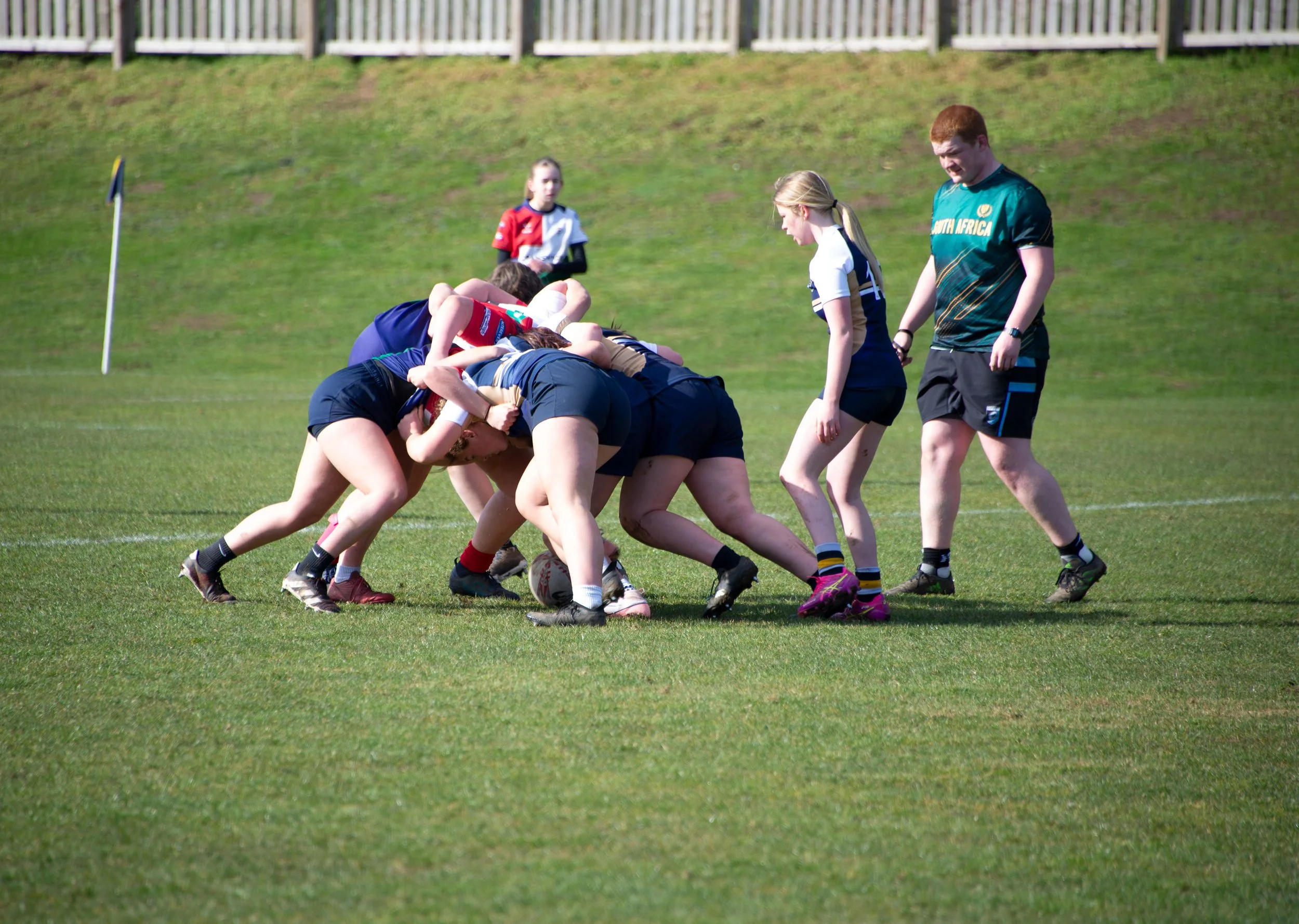 A women's rugby scrum on a green field with players in dark blue, red, and white uniforms; a referee or coach in a teal shirt with 'South Africa' written on it observing nearby.