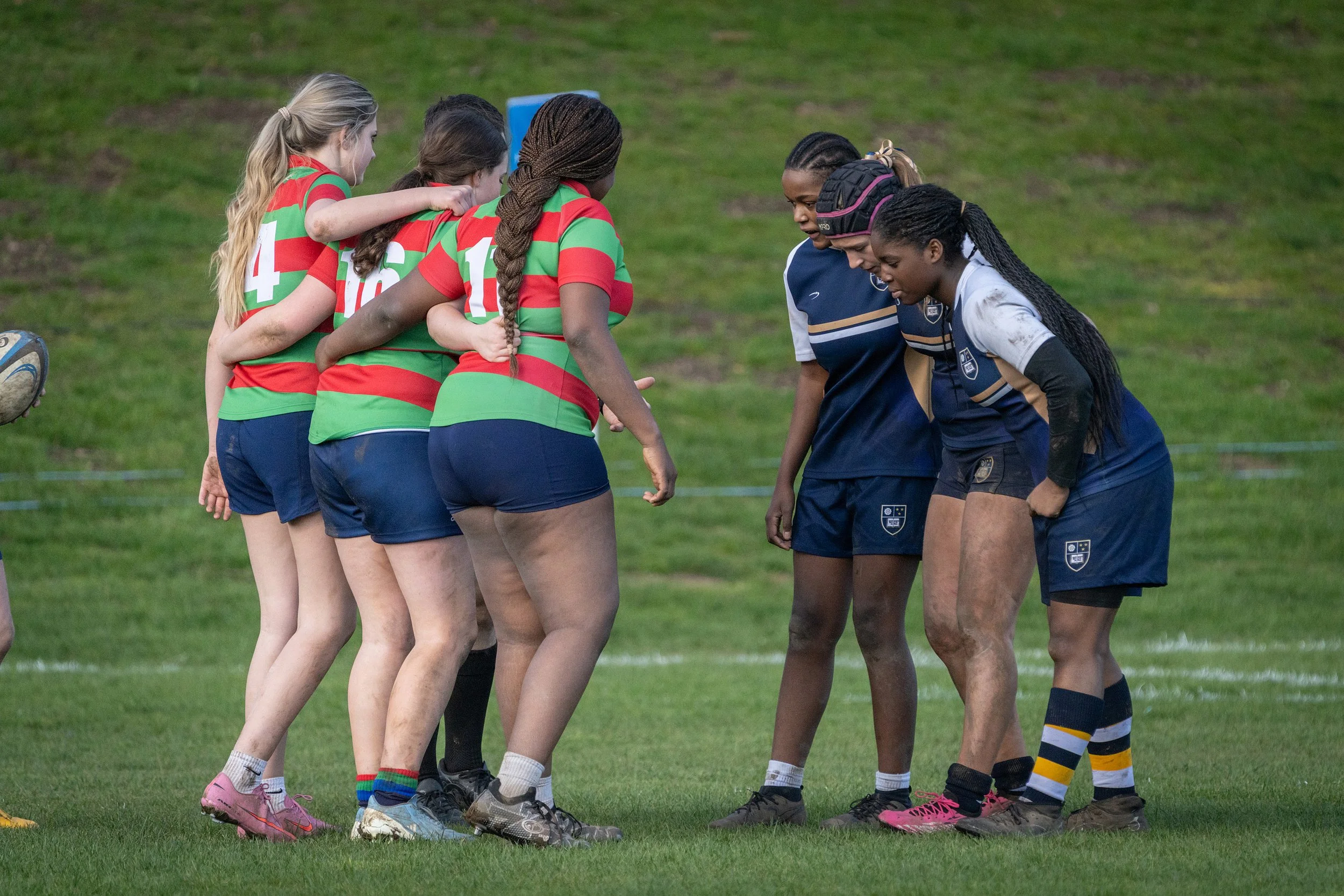 Women rugby players from two teams huddle on a grassy field before a match, with their heads bowed and hands joined in the center.