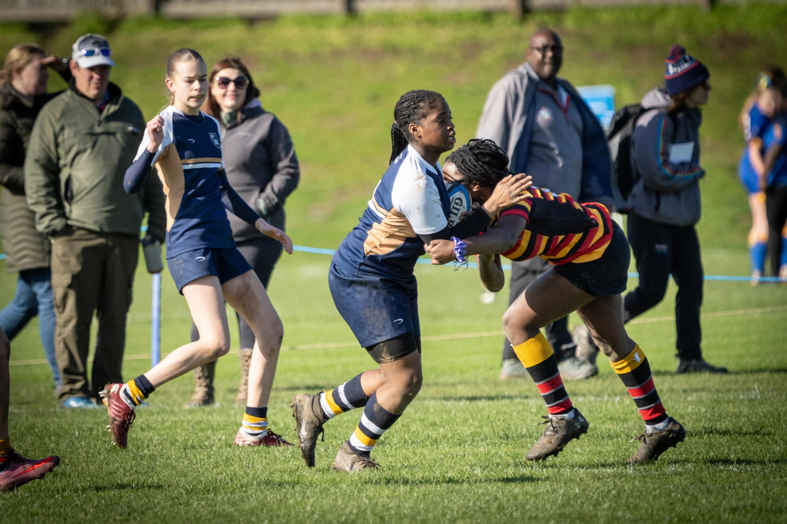 Women playing rugby on a grassy field with spectators watching.