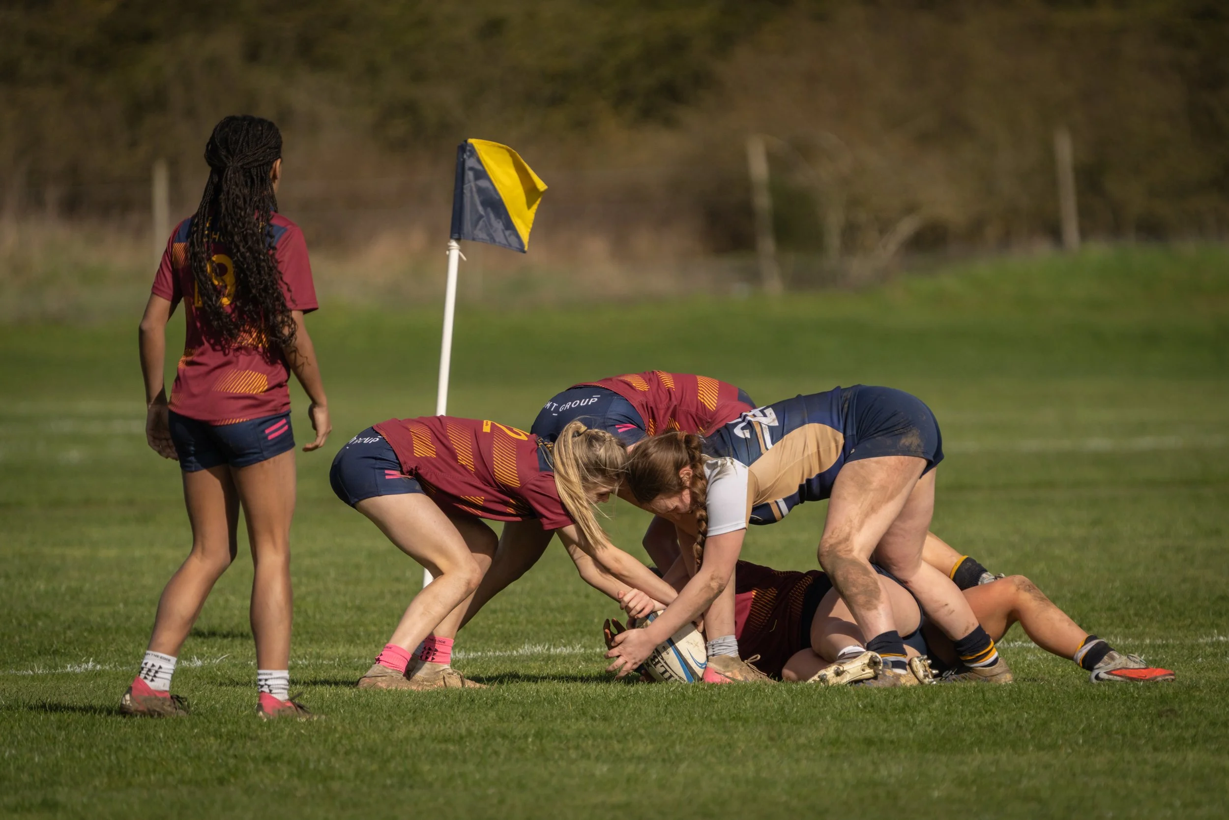 Women rugby players forming a scrum on a grassy field, with a girl standing nearby and a corner flag in the background.