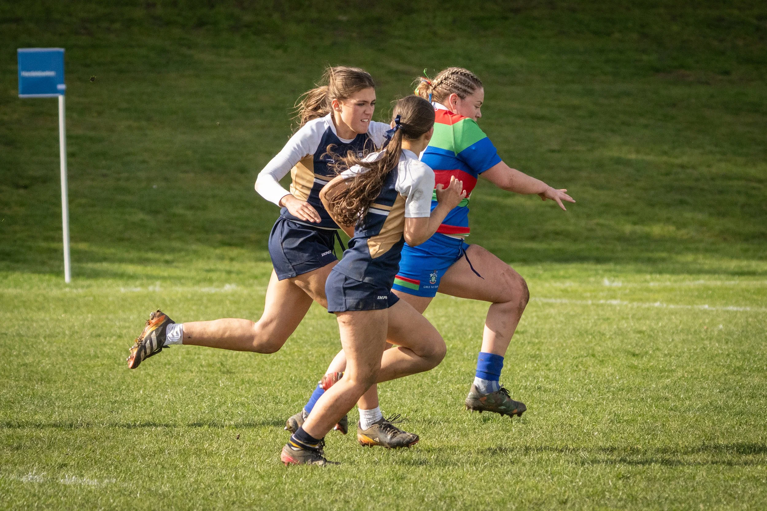 Four young women playing rugby on a grassy field, running in same direction with determined expressions.