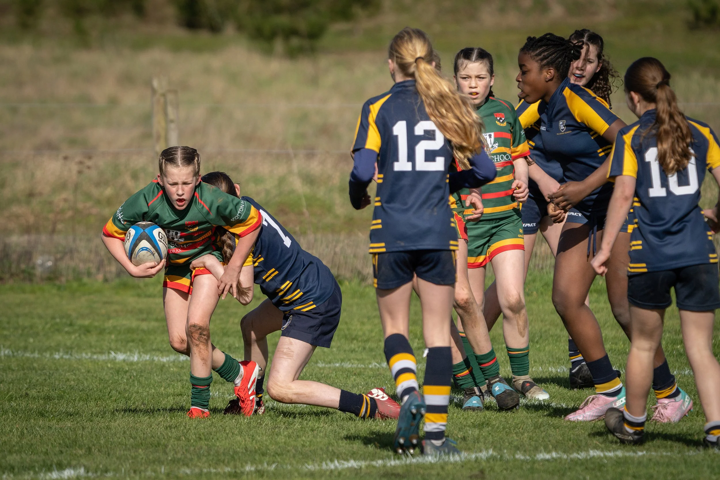 Girls playing rugby on a grassy field, some in green and others in navy blue and yellow uniforms, with one girl holding a rugby ball.