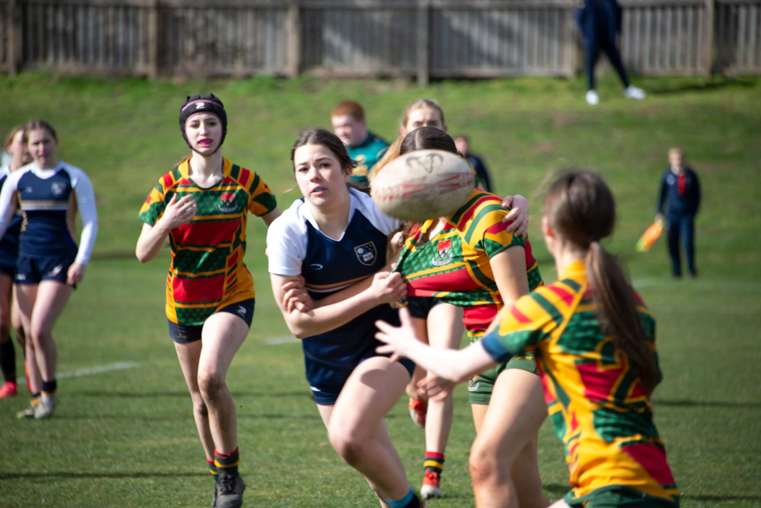 Group of girls playing rugby on a grassy field, wearing colorful uniforms and running towards a rugby ball.