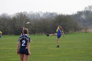 Girls playing rugby on a grassy field, one girl kicking the ball while others watch.
