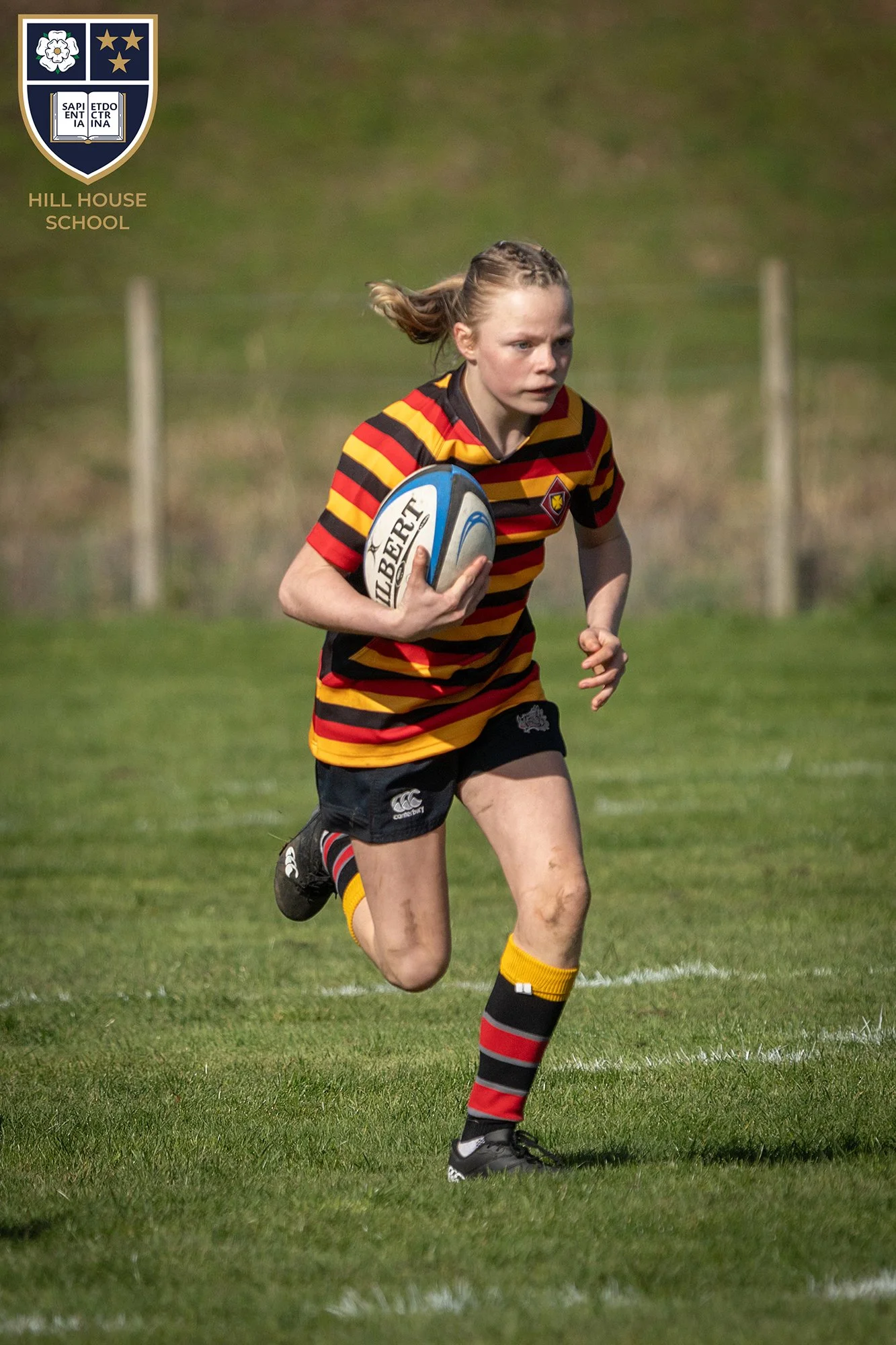 A young female rugby player running on a grass field while holding a rugby ball, wearing a striped red, yellow, and black jersey, black shorts, and matching socks.