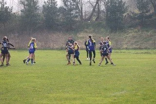 Female rugby players competing on a grassy field, some wearing jerseys with the number 10, others in darker gear, with trees in the background.
